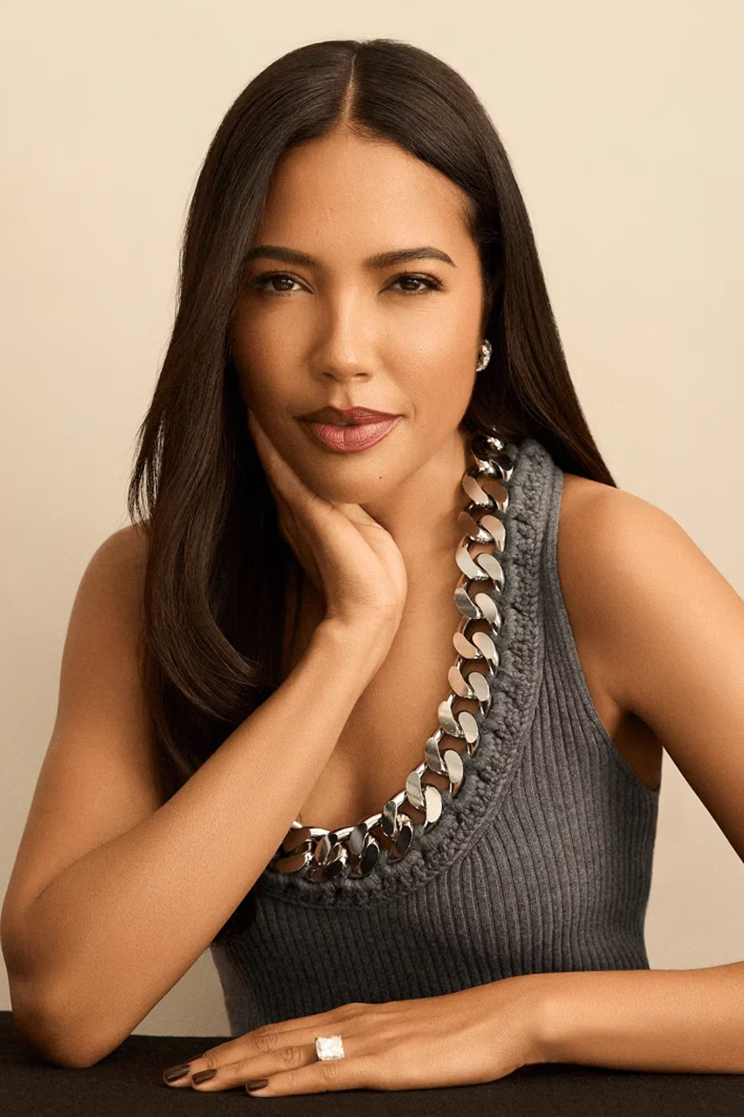 A woman with long dark hair, wearing a sleeveless gray dress with metallic chain details around the neckline, resting her chin on her hand, looking at the camera against a plain beige background.
