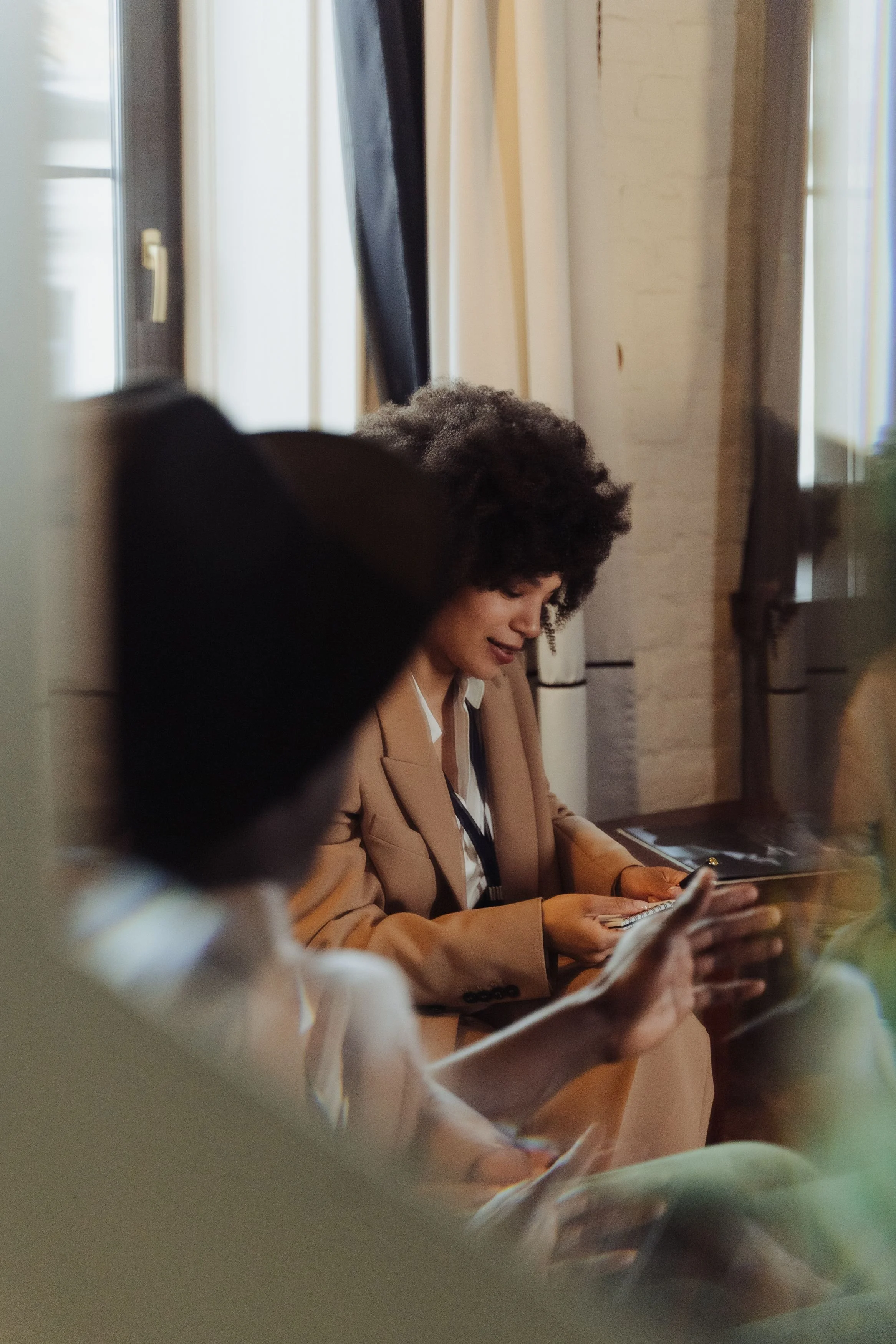 A woman with curly hair wearing a beige blazer is sitting at a table, looking at her phone, with other people partially visible in the foreground and background, in a well-lit room with white brick walls and large windows.