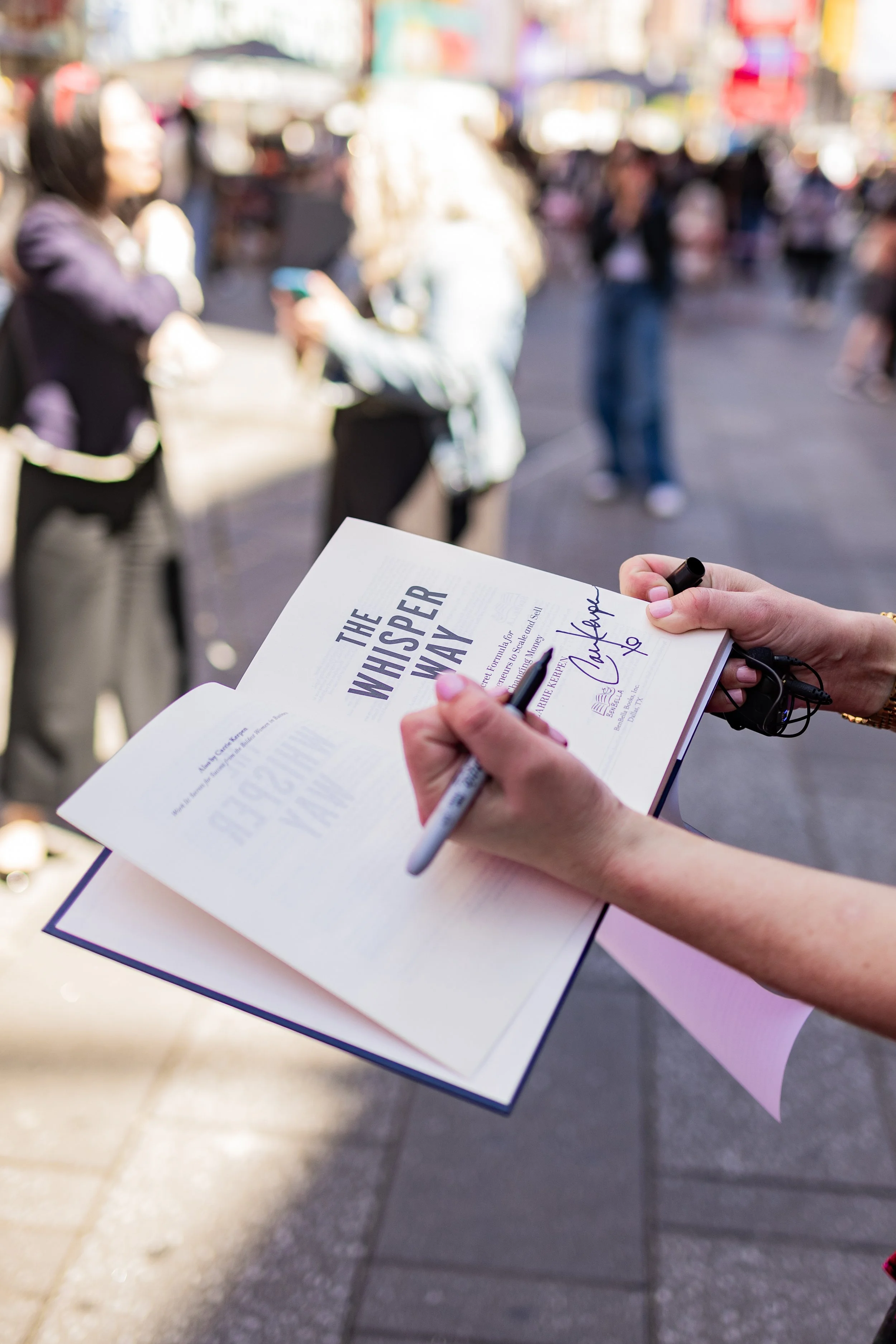 Person holding signed copy of a book titled 'The Whisper Way' at a busy outdoor event or street fair.