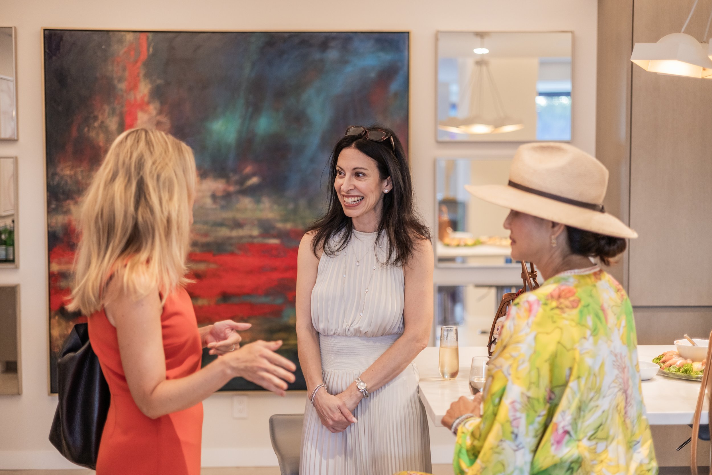 Three women are engaged in conversation at an art gallery or event space. One woman is wearing a coral dress, another a white striped dress with sunglasses on her head, and the third appears in a colorful floral top with a large hat. Behind them, the