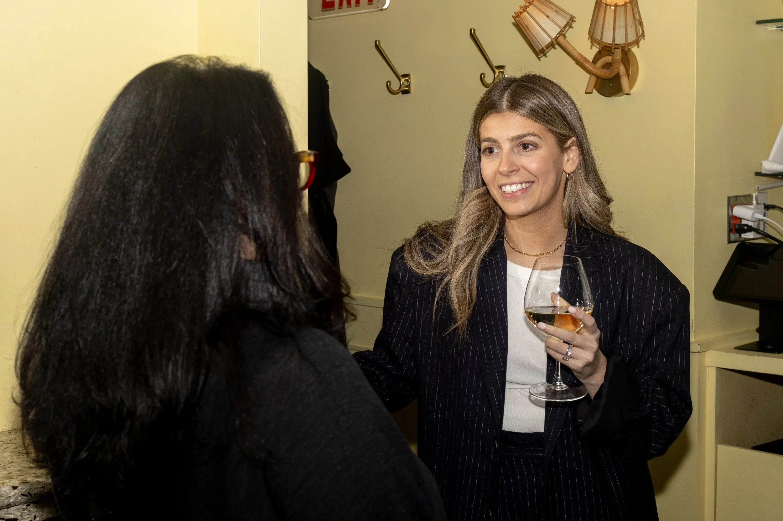 Two women engaging in conversation; one is holding a wine glass, both smiling, in a room with hooks and a wall lamp.