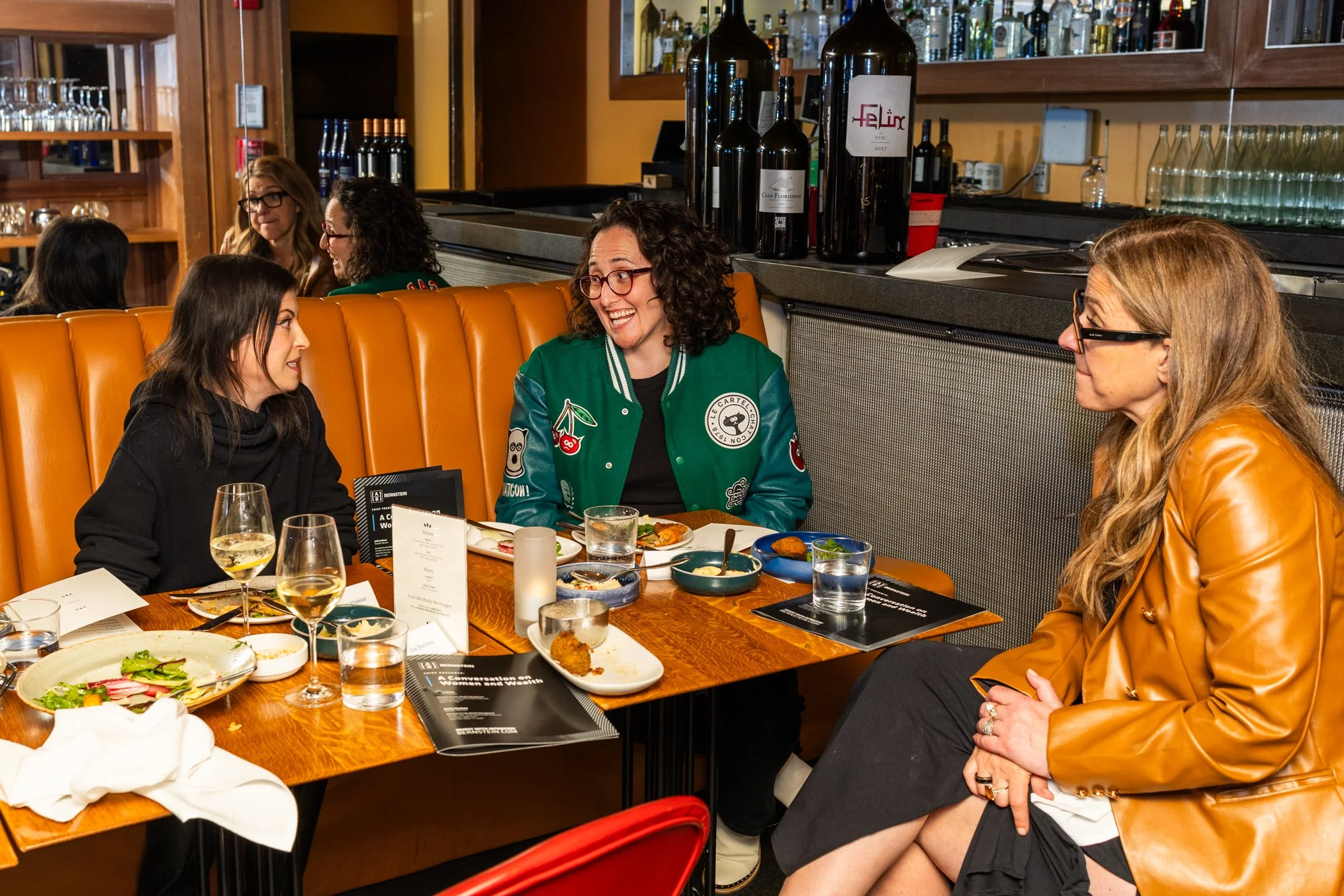 A group of women having a conversation at a restaurant table. The women are smiling and engaging with each other. The table has various plated dishes, drinks, and menus. The background shows shelves with bottles and glasses.