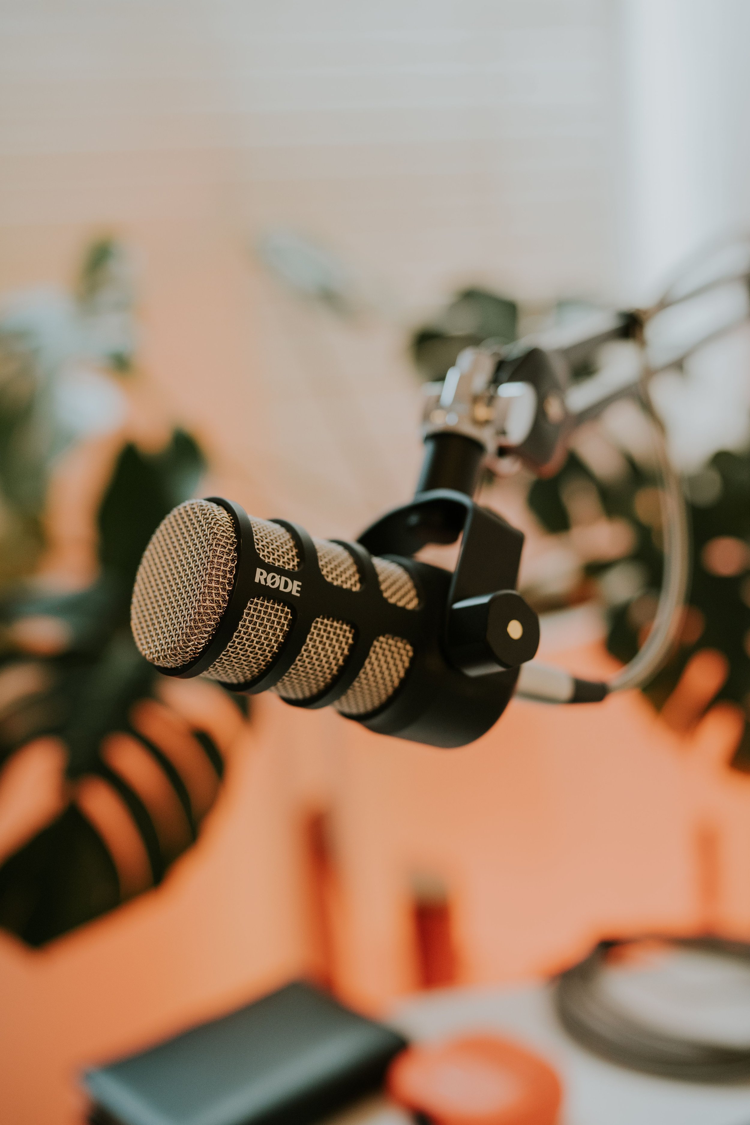Close-up of a beige Rode microphone mounted on a black stand with a hand in the background against a soft orange wall, with blurred objects including a white plate with a piece of orange and other indistinct items.