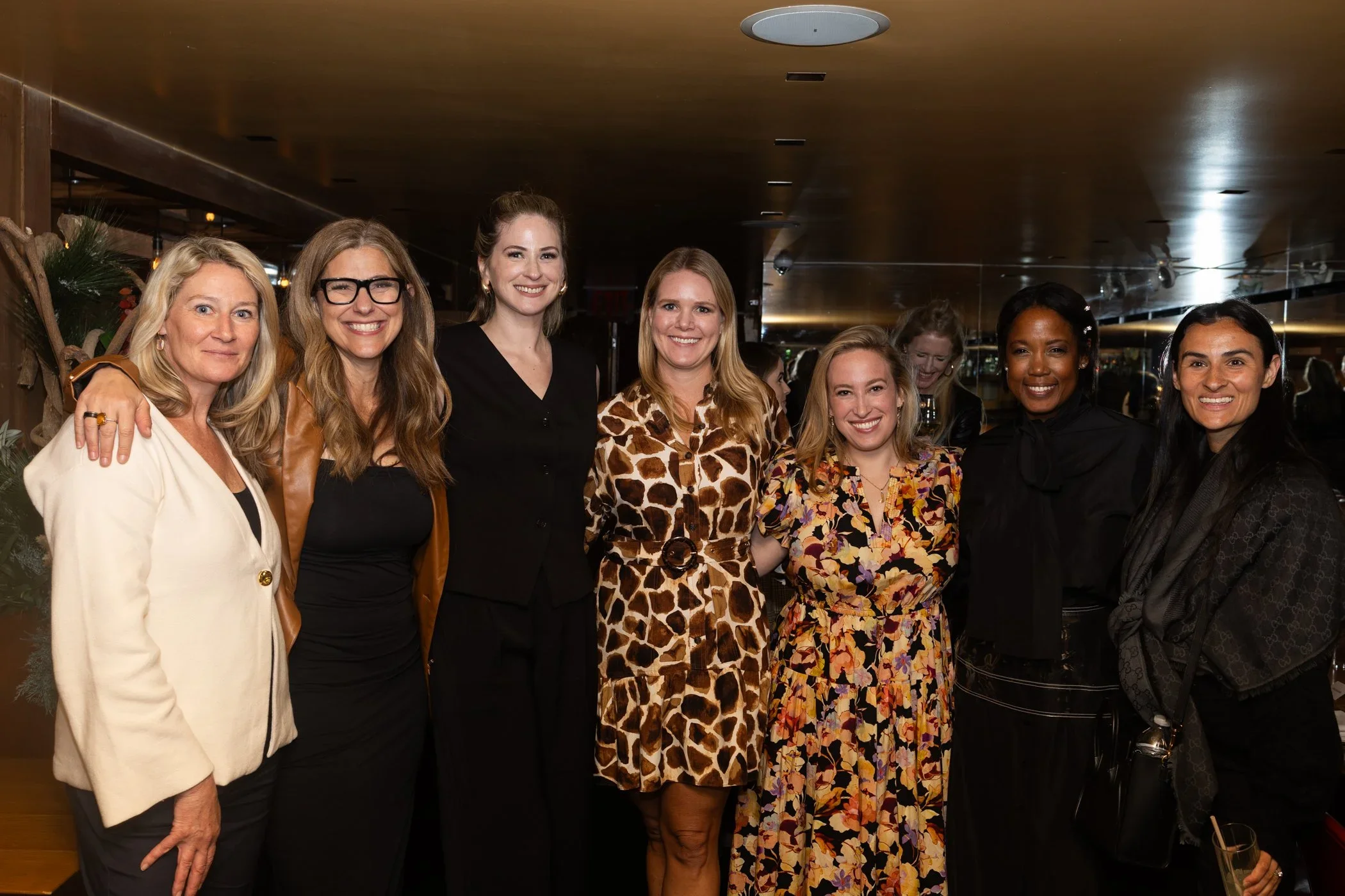 A group of seven women posing together at a social event in a dimly lit venue. They are smiling and dressed in fashionable outfits.