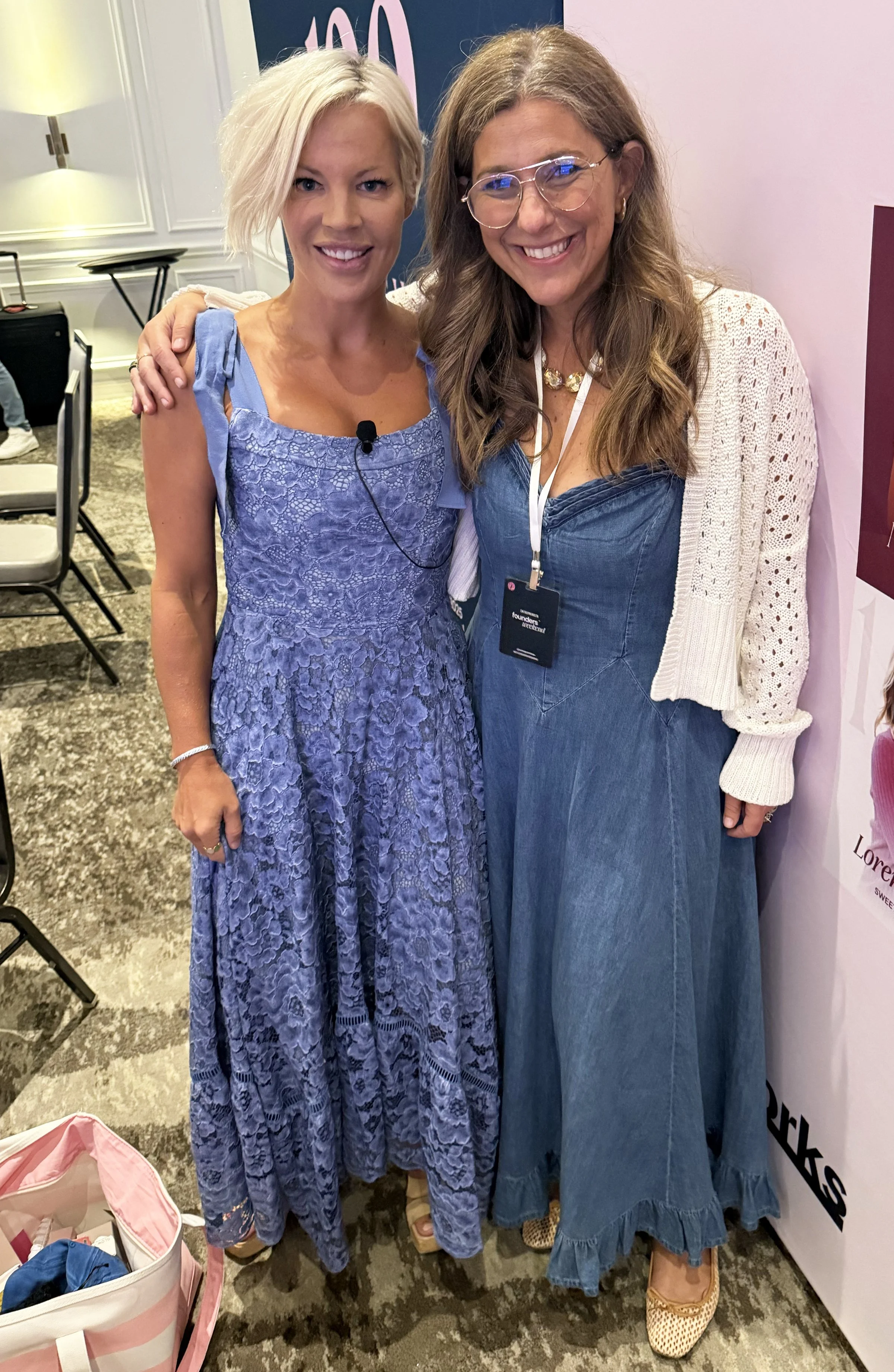 Two women standing close together, smiling at the camera, with one woman wearing a lavender lace dress and the other in a denim dress with a white knit cardigan, in an indoor event room.