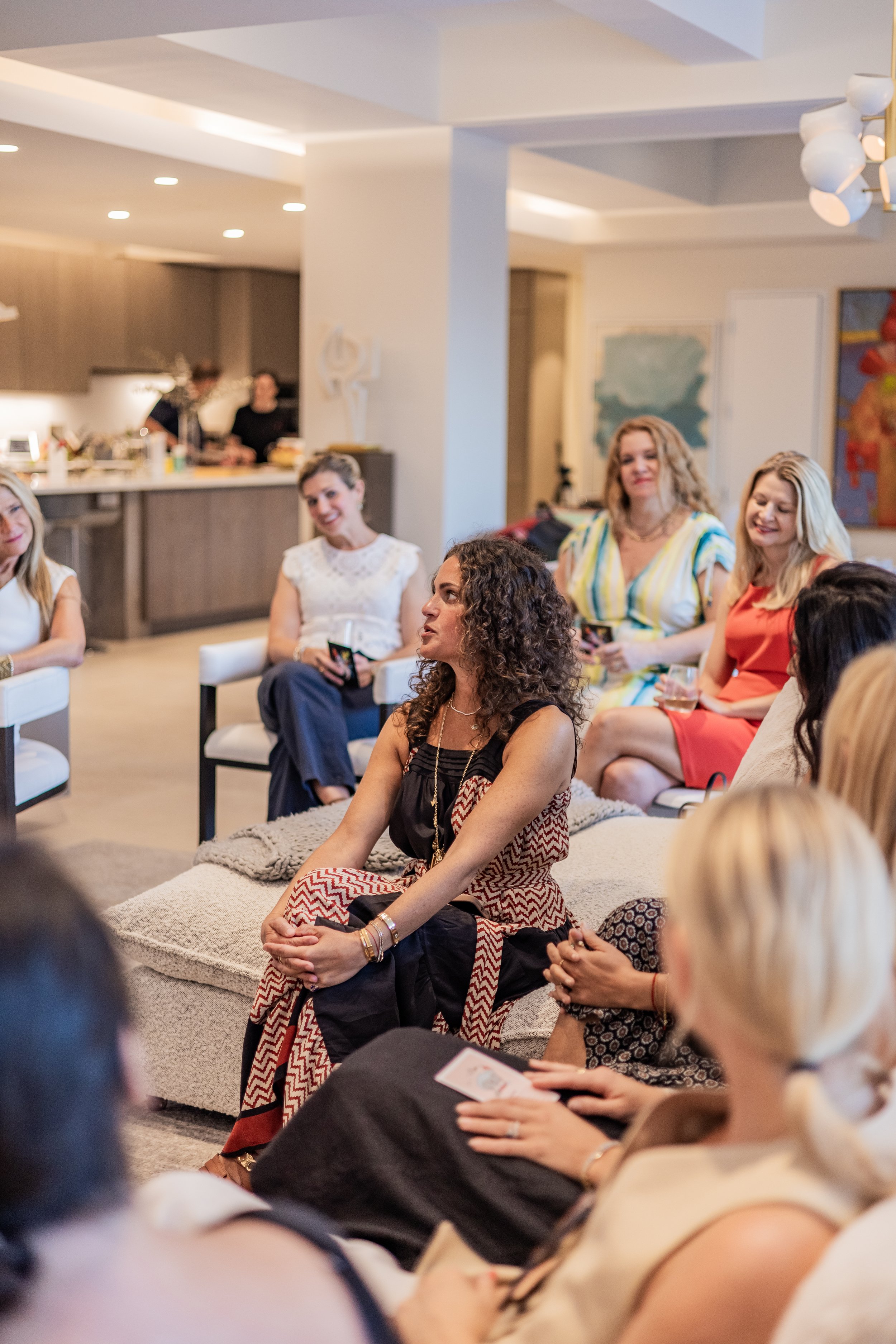 Women seated in a living room, engaging in a social gathering; some are holding drinks and others are looking at their phones.