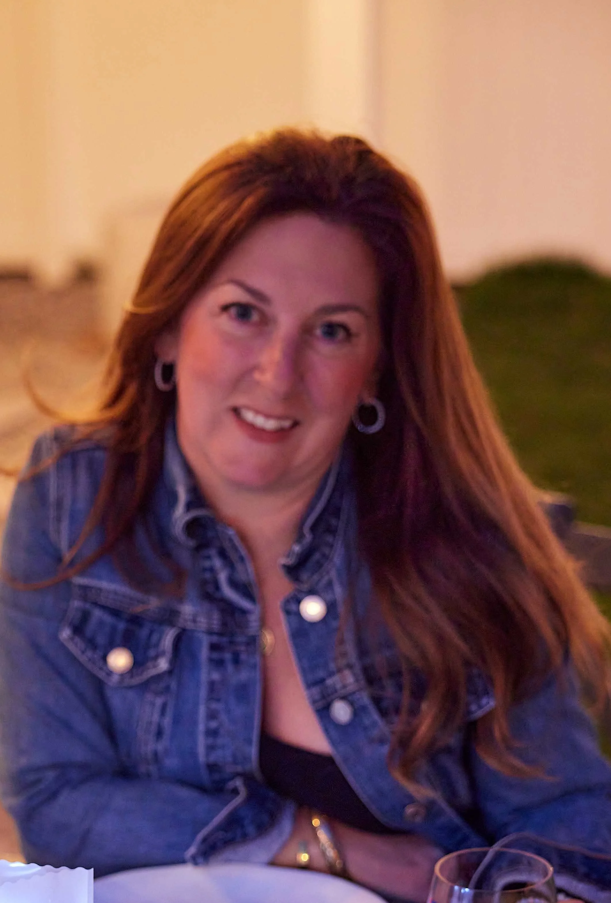 A woman with long red hair wearing a denim jacket sits at a table in a dimly lit room, smiling at the camera. There are glasses on the table in front of her.