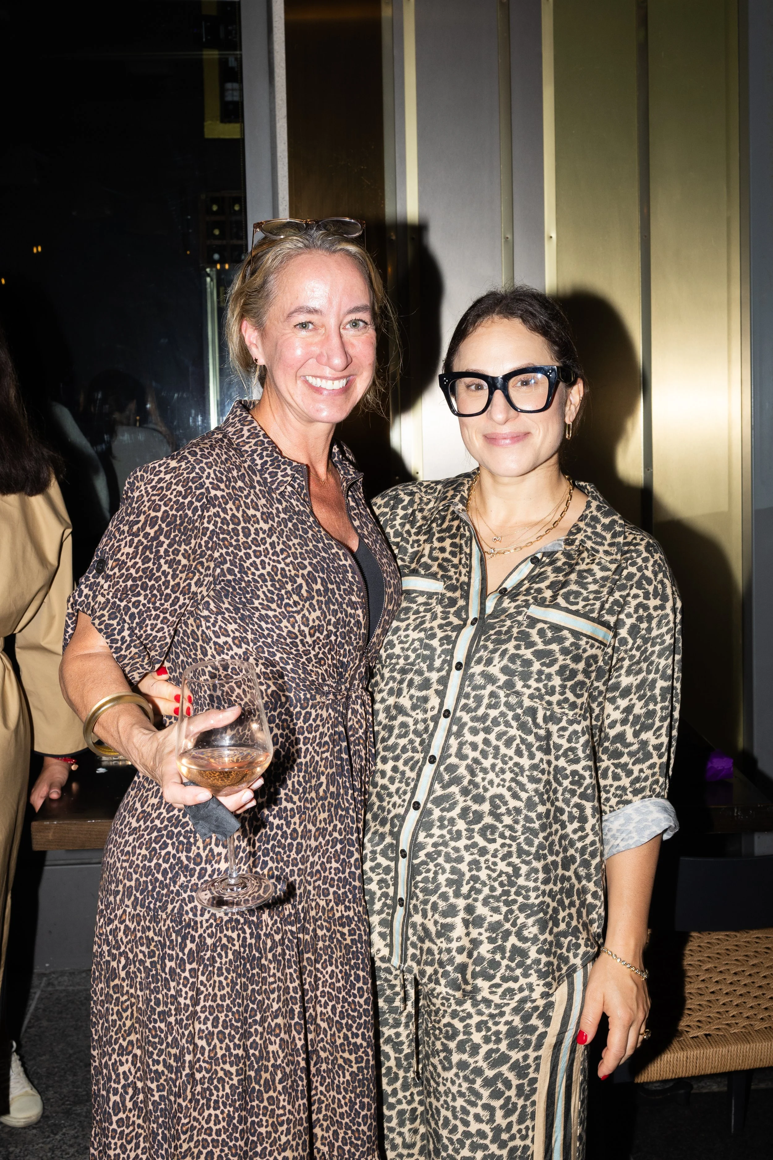 Two women standing close together at an indoor event, both smiling at the camera. The woman on the left has blonde hair, is wearing a leopard print dress, and is holding a glass of rosé wine in her right hand. The woman on the right has dark hair, glasses, and is dressed in a matching leopard print shirt with a striped panel detail.