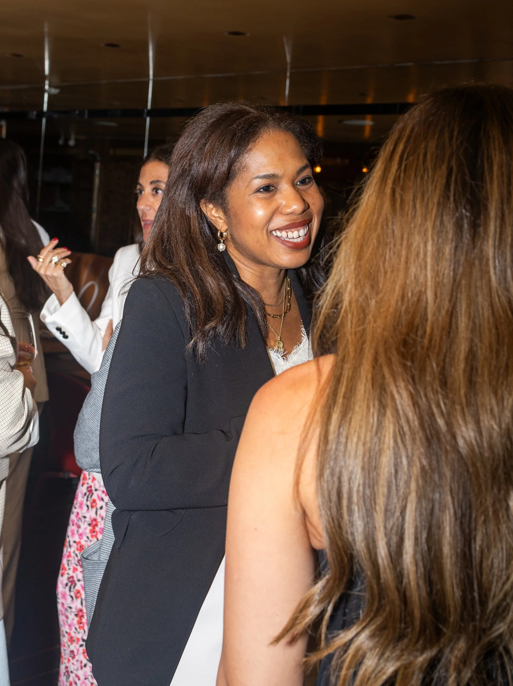 Three women talking at an indoor social event, one smiling and wearing a black blazer with jewelry, the other two partially visible in the background.