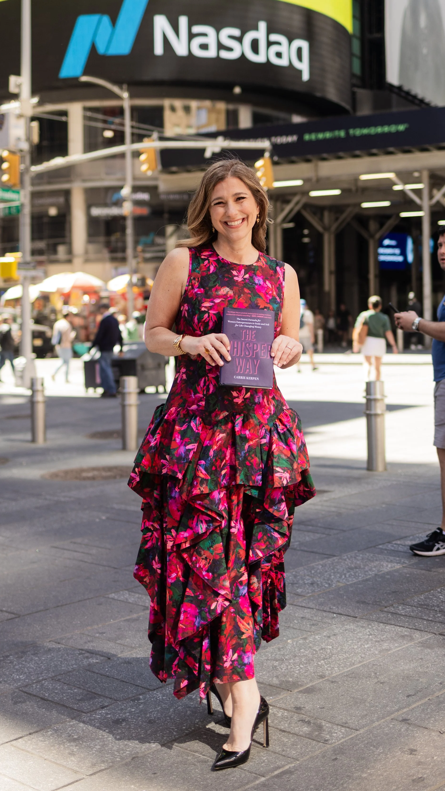 A woman in a vibrant pink, red, and green floral dress walking in Times Square, New York City, holding a book titled 'The Whisper Way' by Carrie Kerpen. She is smiling and wearing black high heels, with a Nasdaq sign in the background.