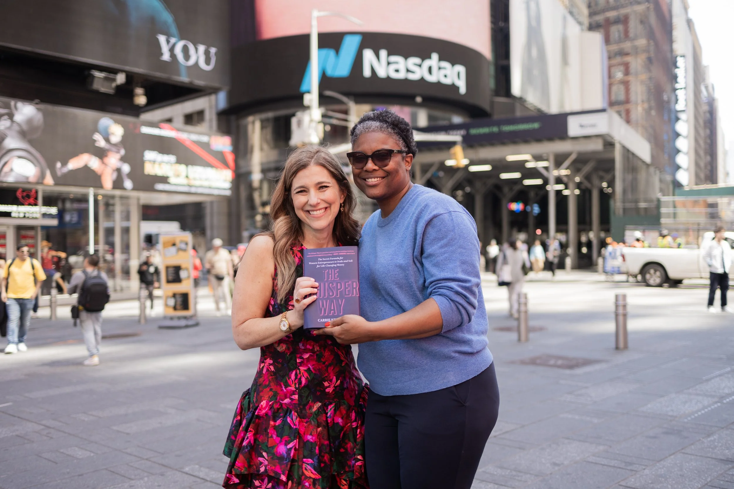 A woman wearing a floral dress and a woman wearing sunglasses and a blue shirt, smiling and holding a book titled 'The Whisper Way' in Times Square, New York City, with NASDAQ sign in the background and busy street scene.