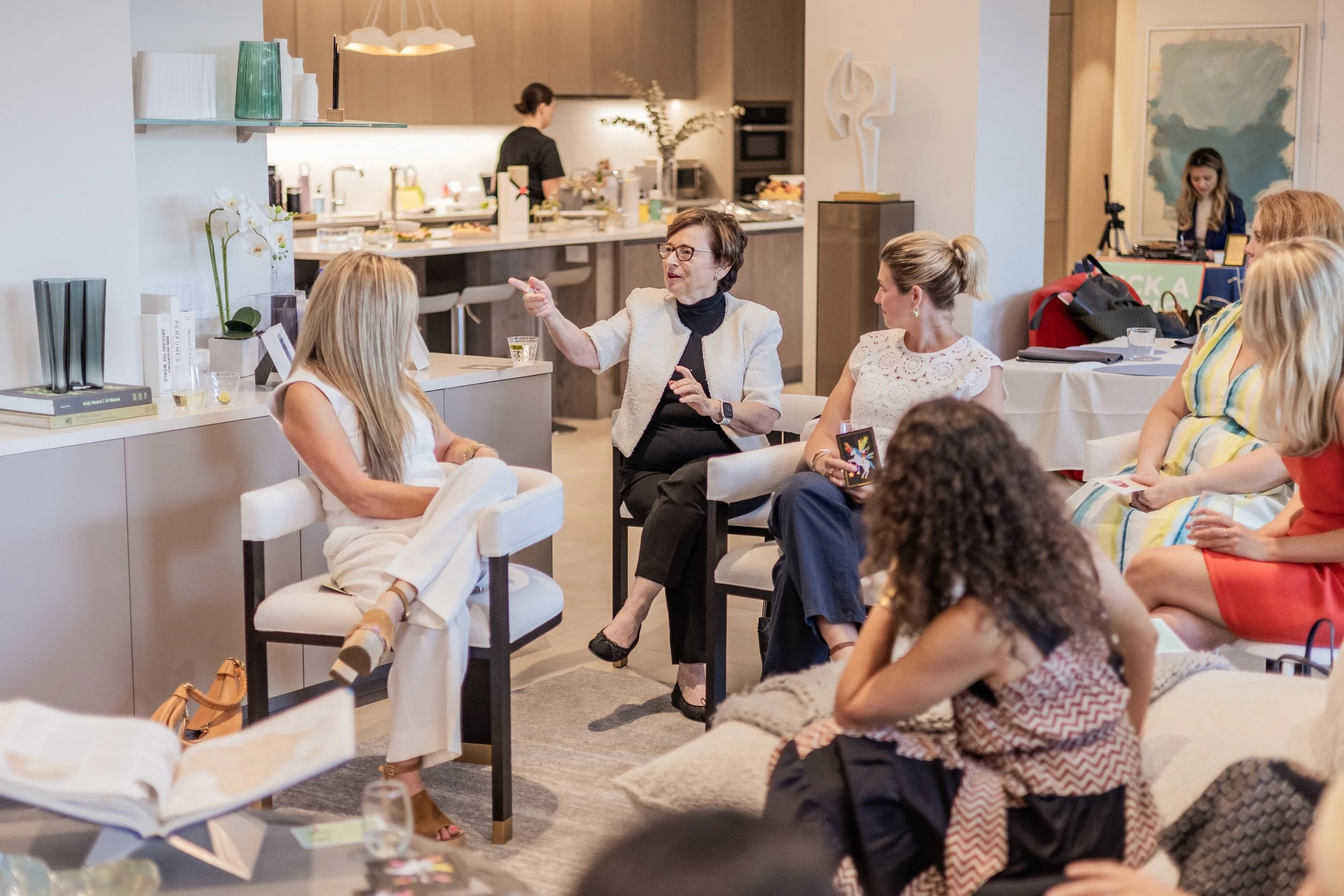 A woman with glasses and dark hair, wearing a light-colored blazer, is speaking or gesturing during a discussion with several women seated around her in a bright, modern interior space. In the background, a woman is working in a kitchen area and another woman is at a table with a camera set up nearby.
