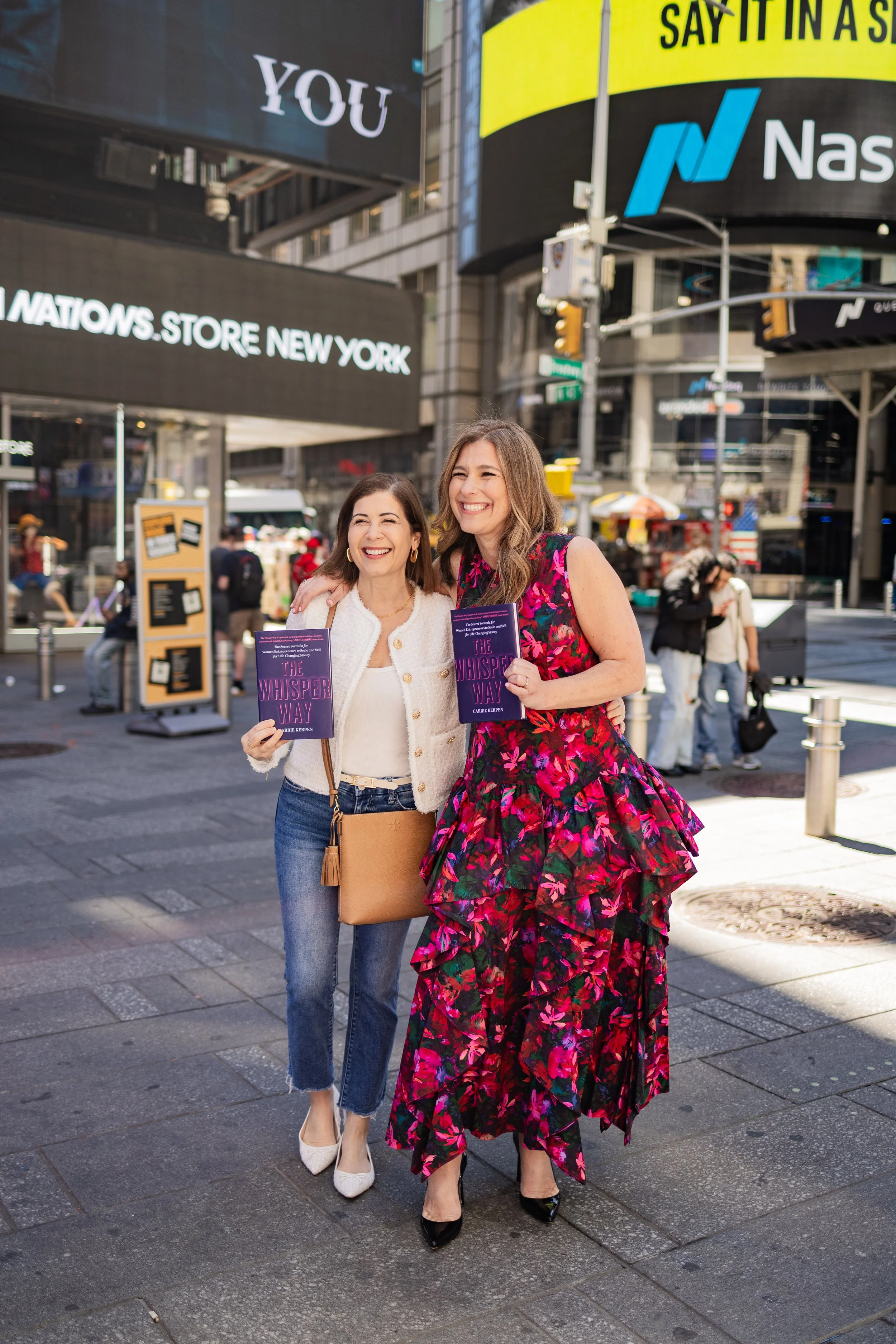 Two women smiling and holding books titled "The Whisper Way" in Times Square, New York City, with digital billboards and city street in the background. One woman is wearing a floral red dress and black high heels, the other is dressed in a white jack