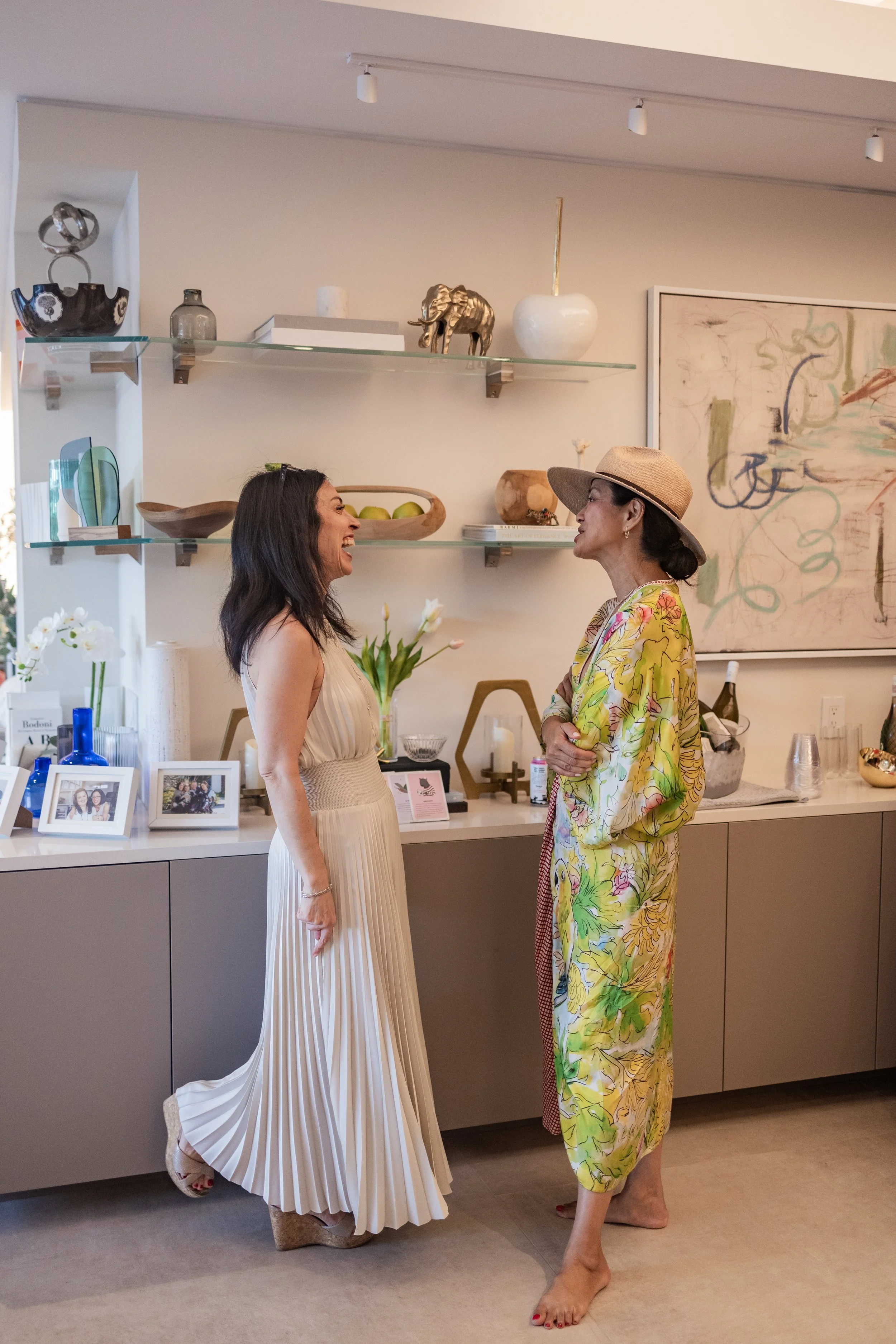 Two women are standing and smiling at each other in a living room or art gallery. One woman is wearing a white pleated dress with high heels, and the other woman is barefoot wearing a colorful floral dress and a straw hat. The background features a wall with decorative objects on glass shelves, framed photographs, and an abstract painting.