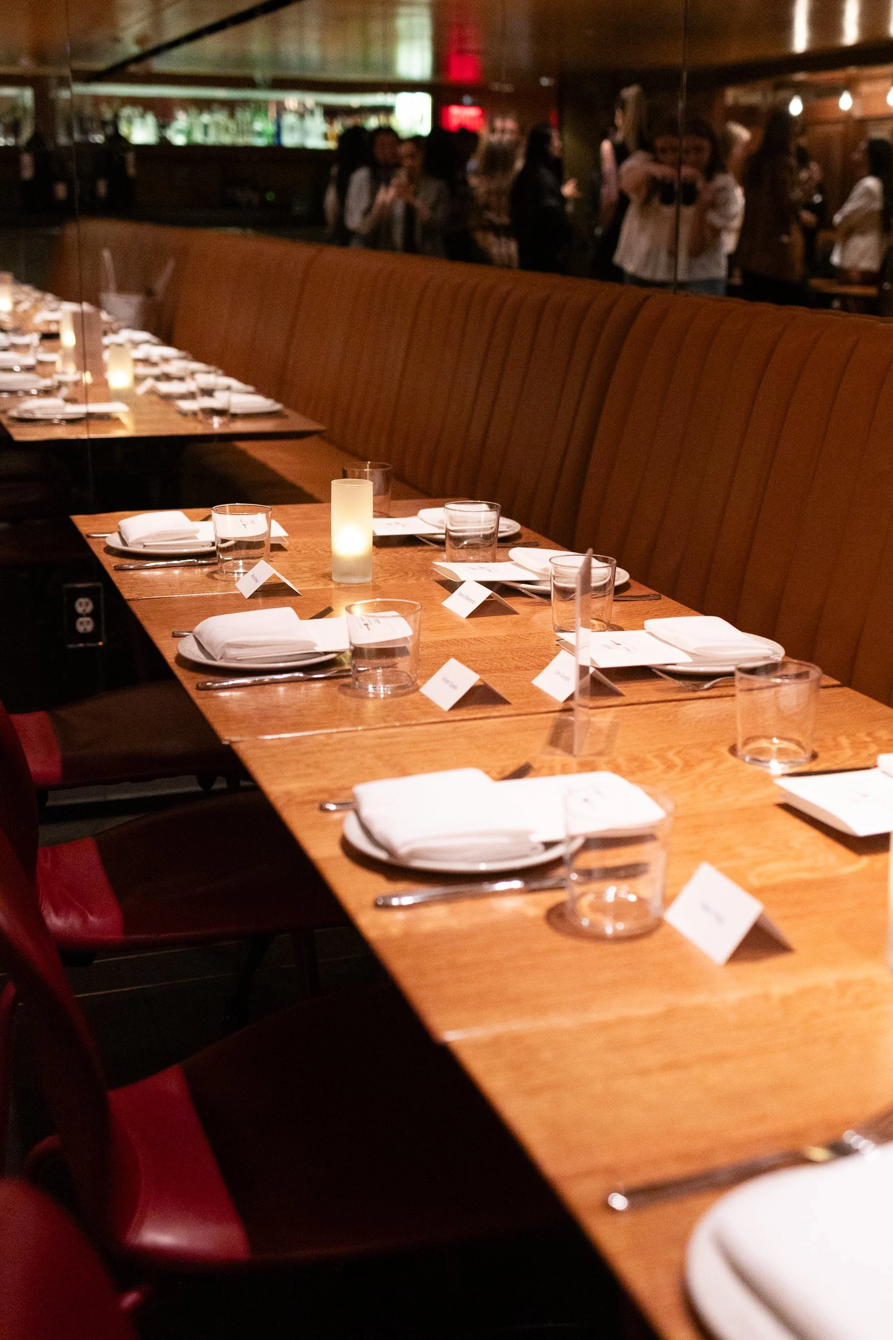 Empty restaurant table with white napkins, glasses, and candles, and a long brown leather banquette against a mirrored wall, with a blurred group of people in the background.