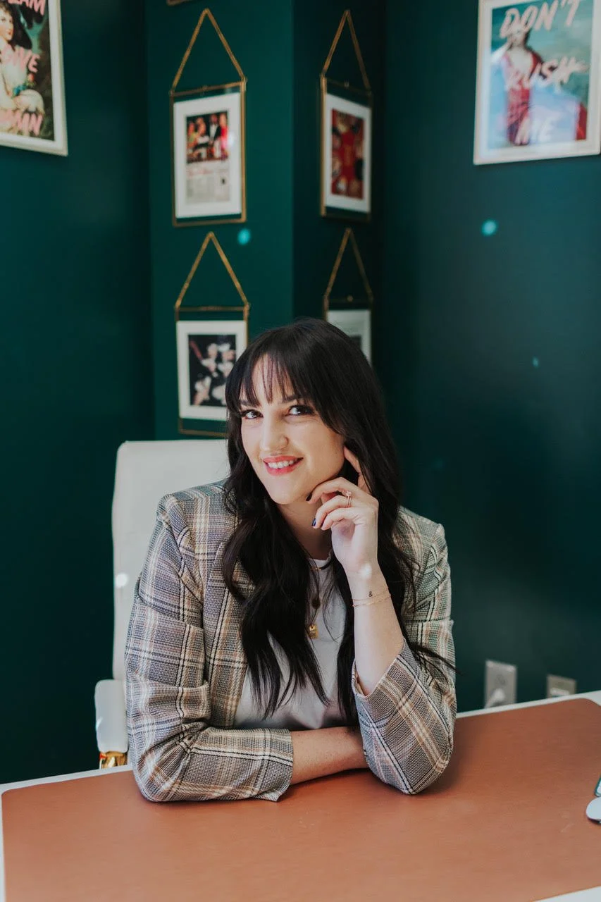 A woman with long dark hair and bangs, wearing a plaid blazer, seated at a desk with a confident smile, in a room with dark green walls decorated with framed pictures.