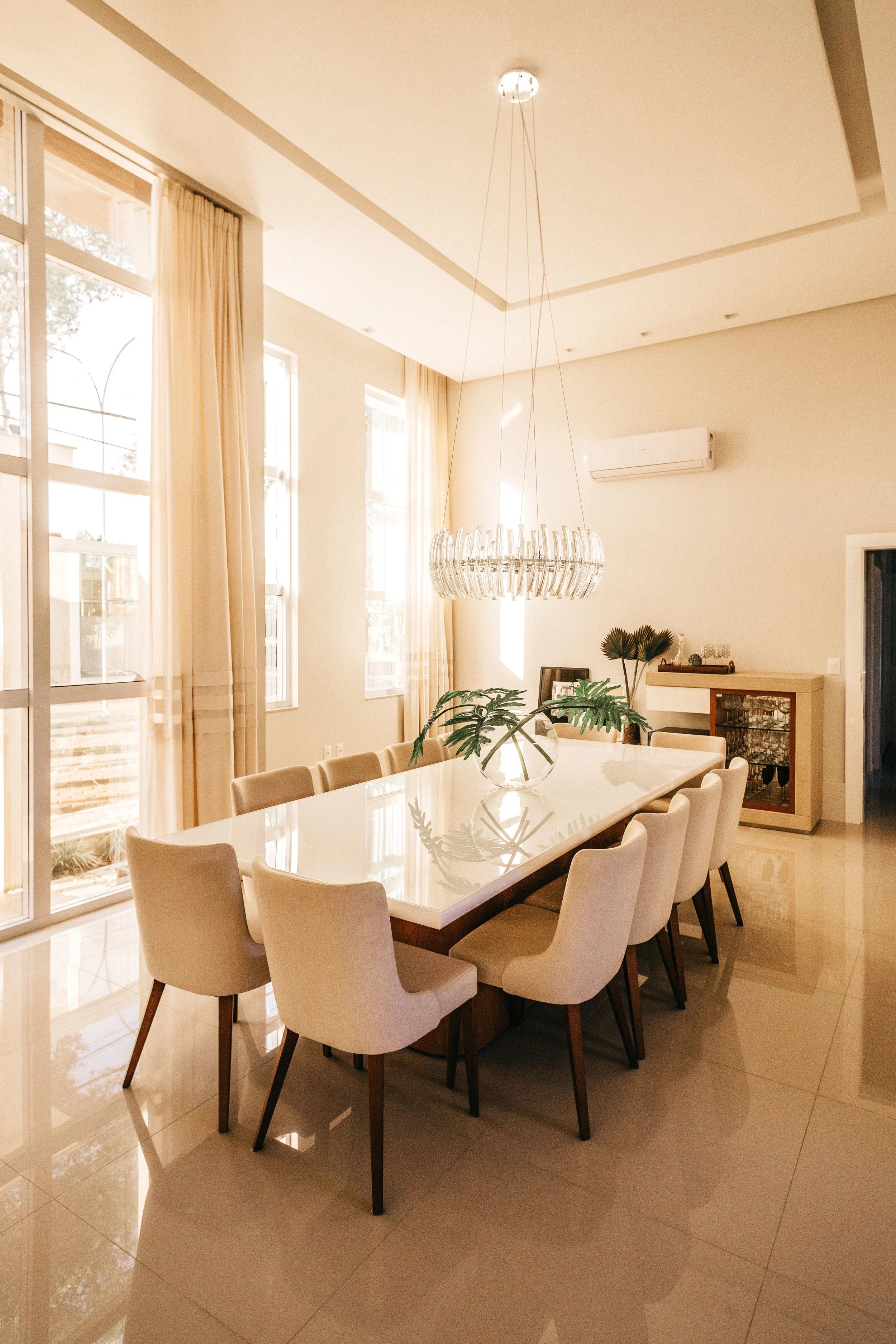 Modern dining room with a white rectangular table, surrounded by beige upholstered chairs, large windows with sheer curtains allowing sunlight, a decorative chandelier hanging above, a side cabinet with plants and decorative items, a neutral color palette, and tiled flooring.