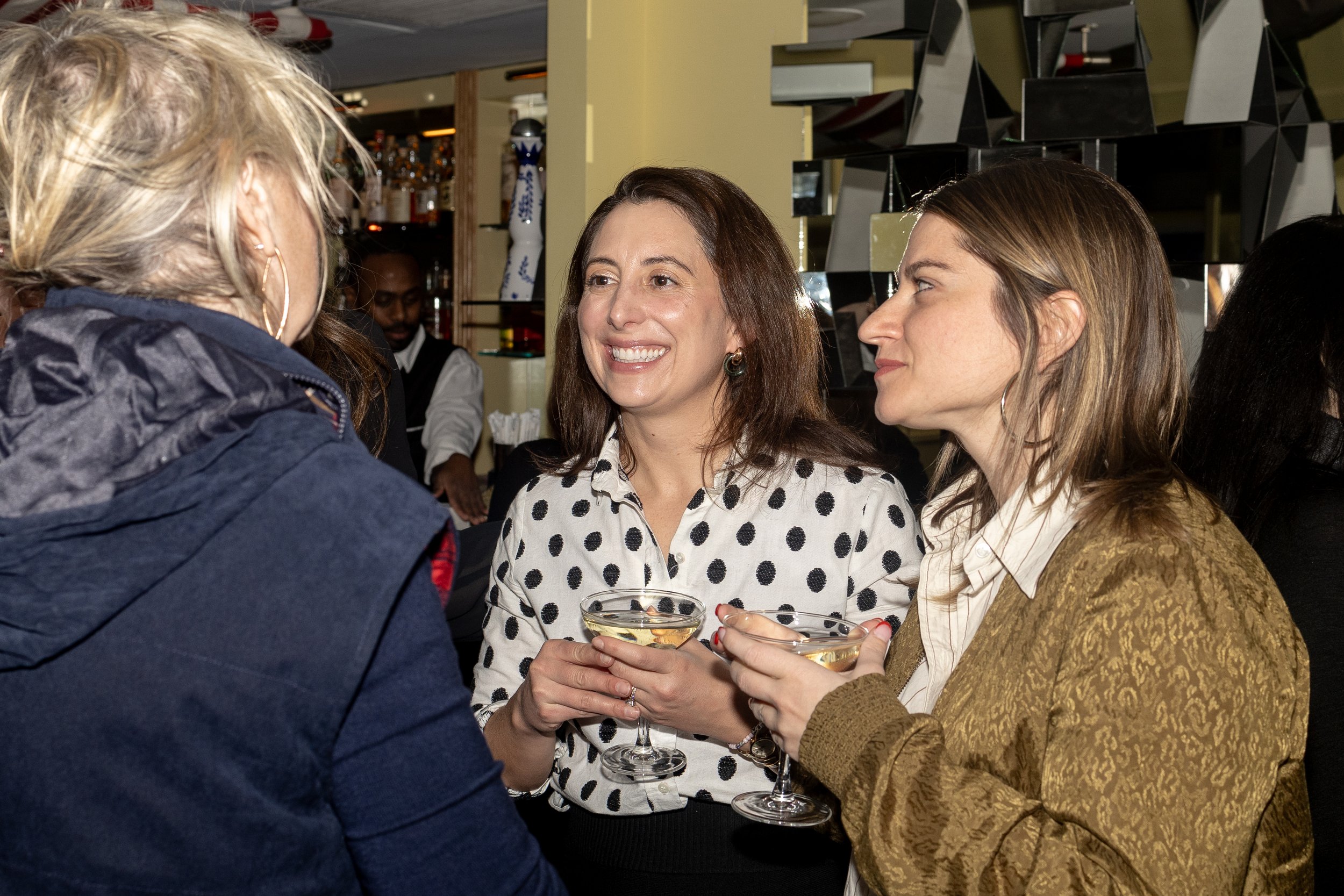 Three women and one man sitting together at a bar, smiling and holding cocktail glasses, engaged in conversation.