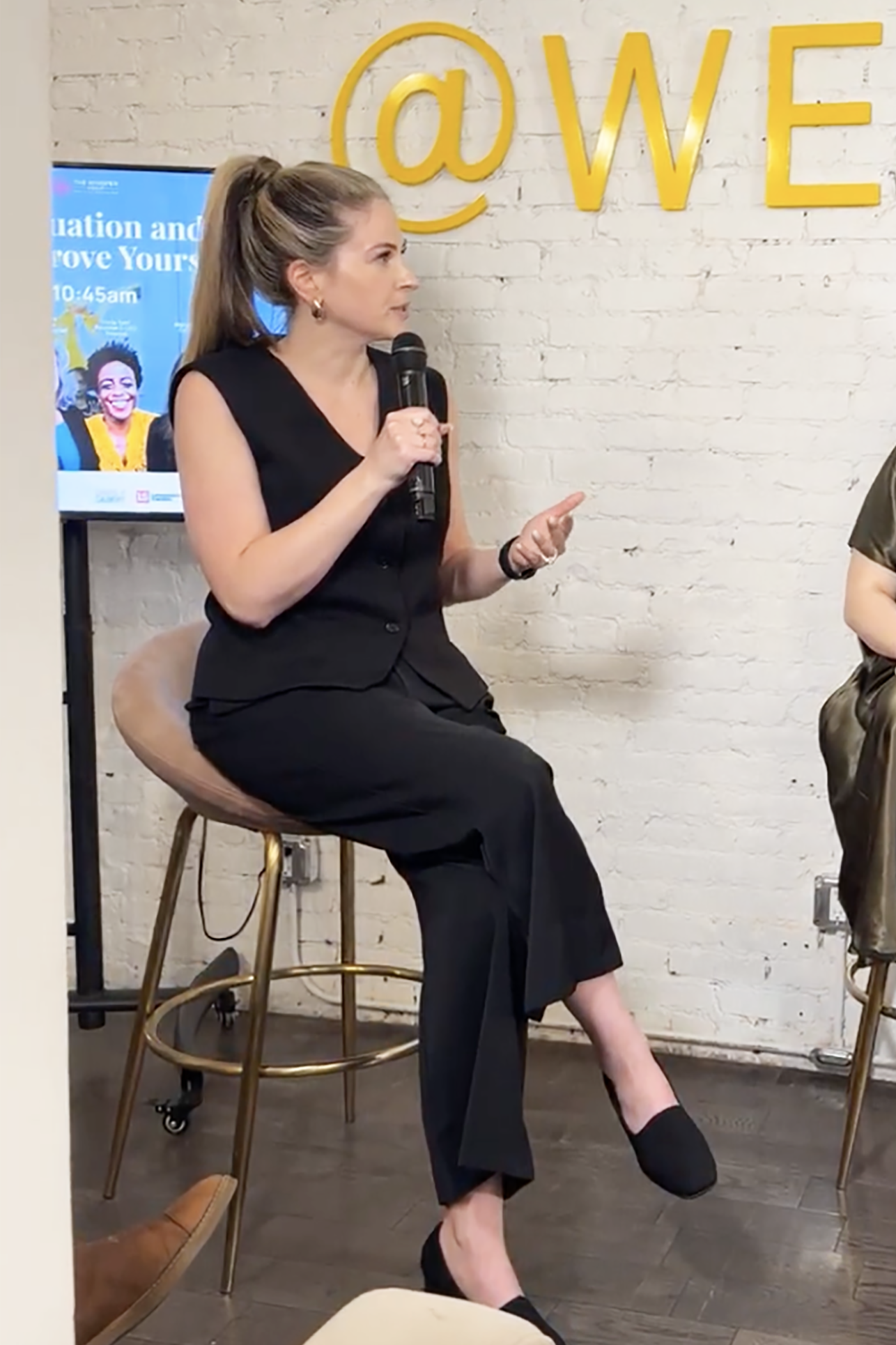 A woman in black dress and high heels sitting on a stool, holding a microphone, speaking in front of a white brick wall with a large gold '@' symbol and the word 'WE'. A digital screen in the background displays a woman smiling, with text about education and a time of 10:45 am.