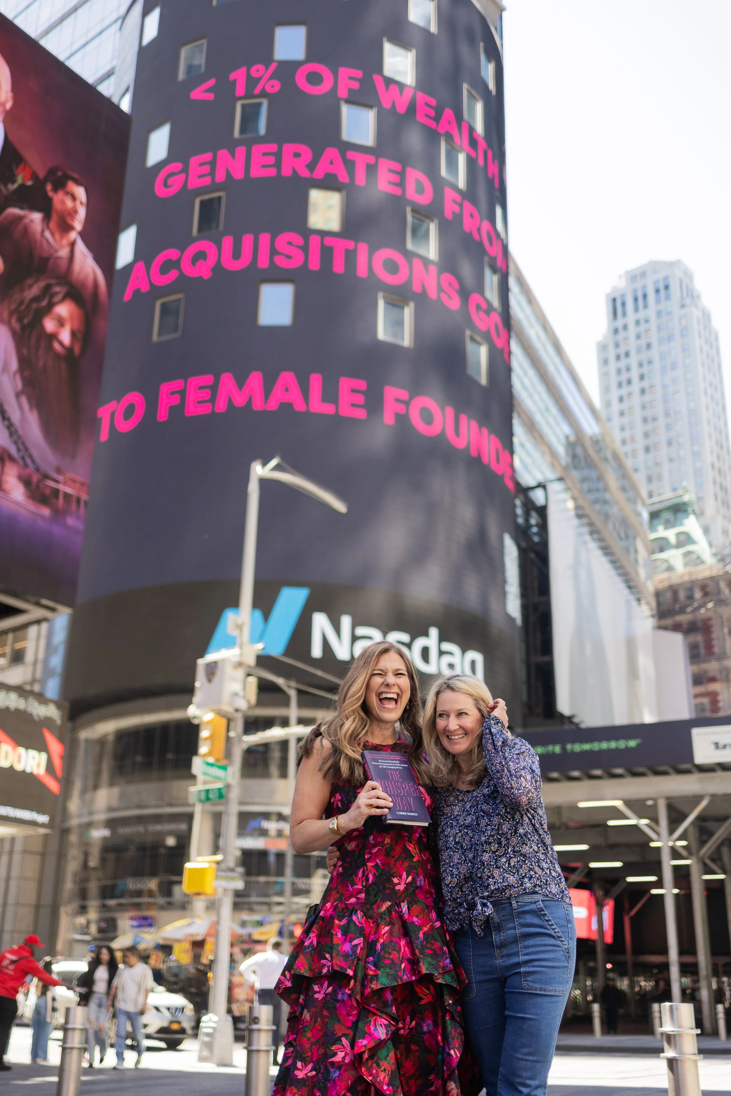 Two women smiling and posing together in Times Square, New York City. One woman is holding a book titled 'The Whisper Way'. There is a large digital billboard behind them displaying text about wealth generated from female founders, and surrounding bu