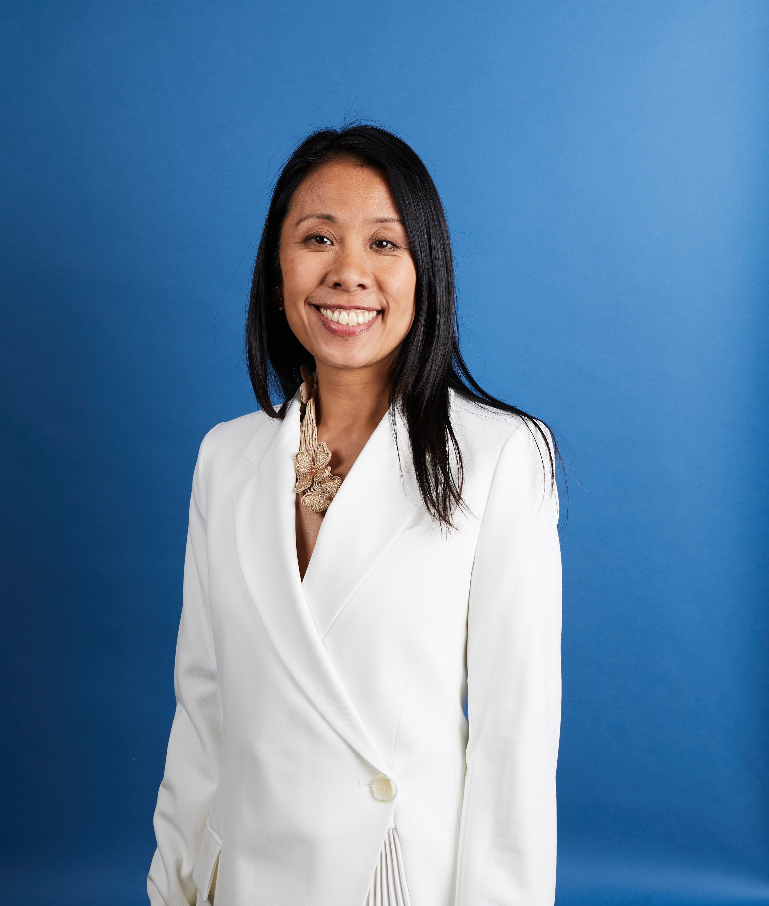 A woman with long black hair, wearing a white blazer and gold necklace, smiling against a blue background.