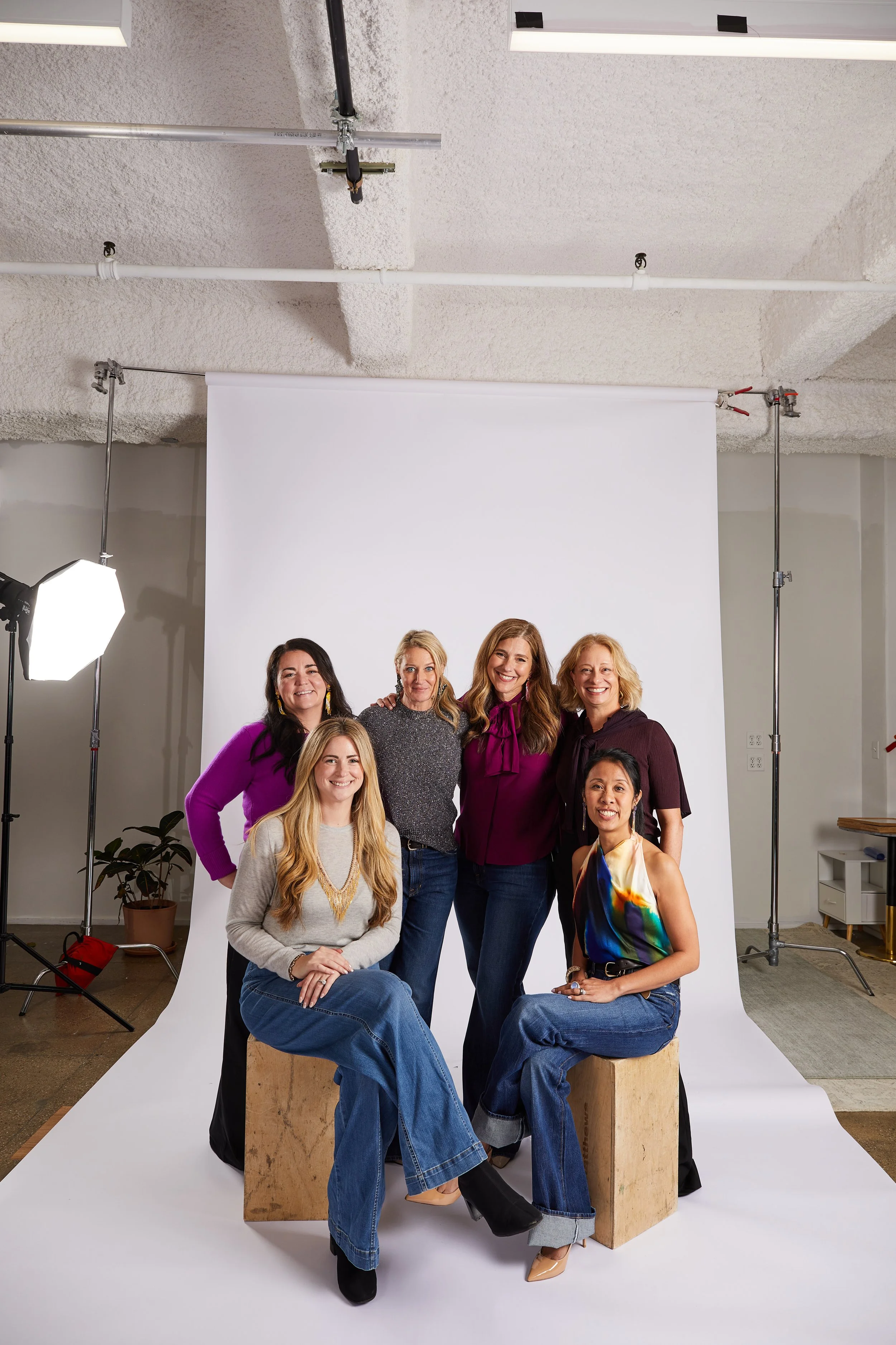 A group of seven women posing together in a photography studio with a white backdrop, professional lighting, and studio equipment visible.