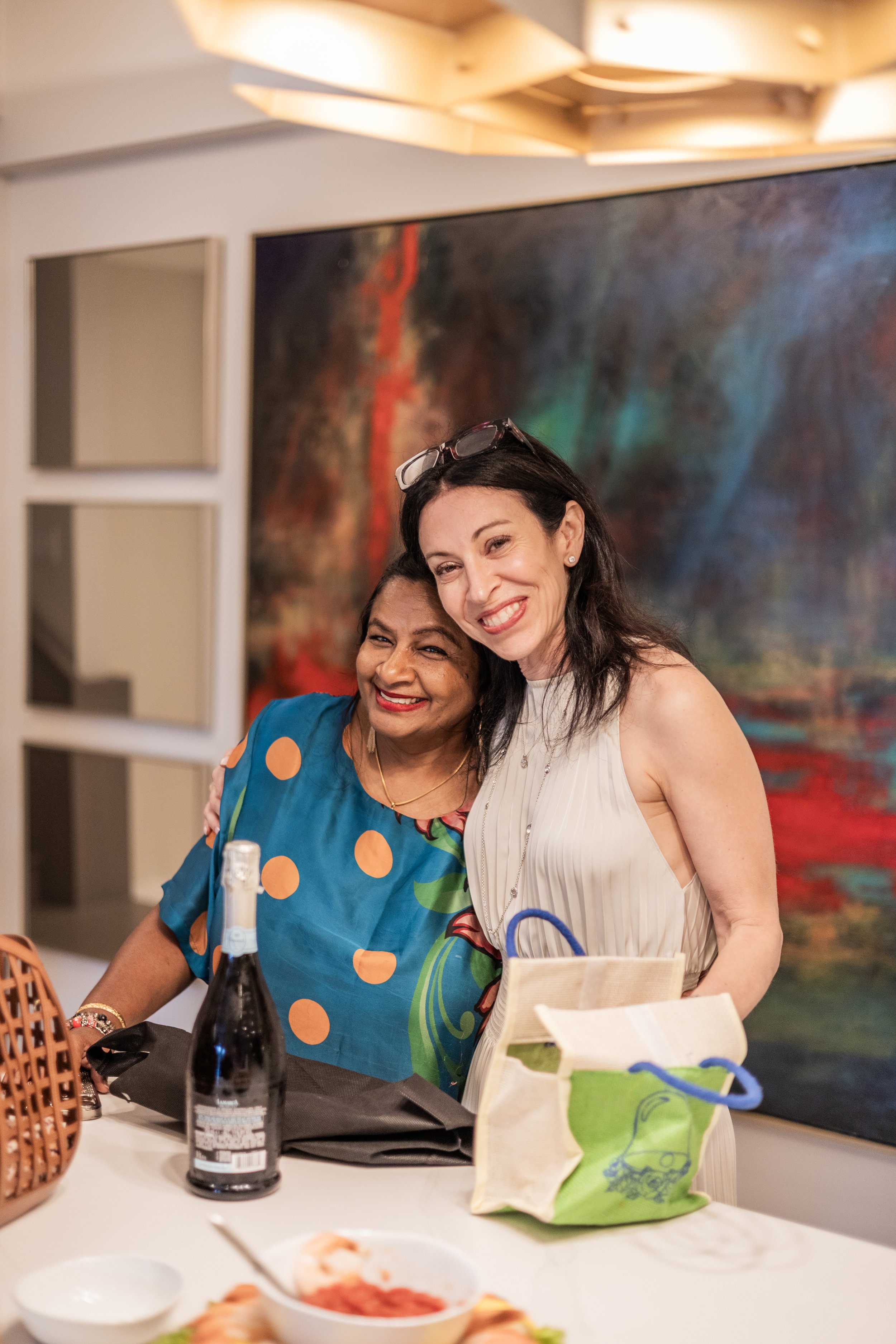 Two women are smiling and posing for a photo in a restaurant or party setting. One woman is wearing a blue dress with orange polka dots, and the other is wearing a white sleeveless top. They are standing behind a table with food, drinks, and a gift b