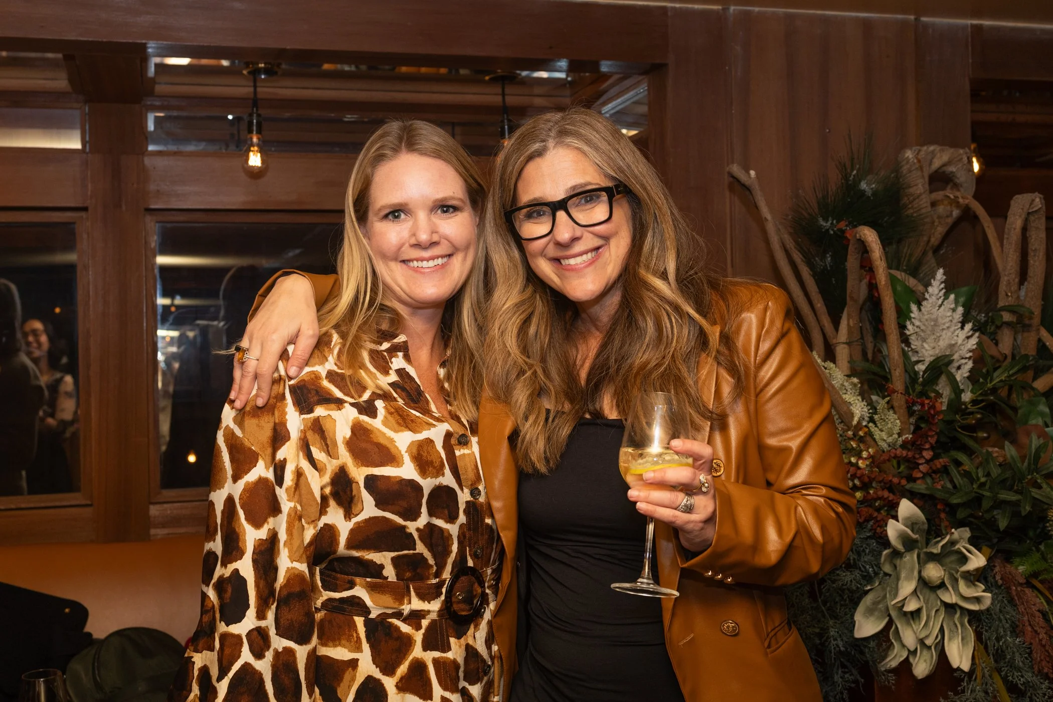 Two women smiling and posing together indoors at night; one wearing a giraffe print dress, the other in a brown leather jacket holding a drink with lemon.