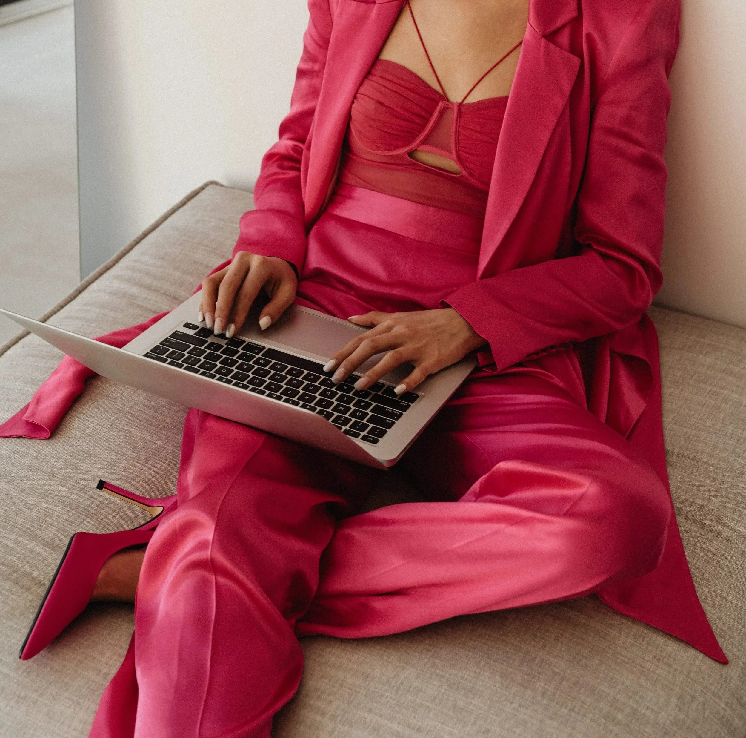Woman wearing a red jacket and matching red dress, sitting on a beige sofa, typing on a silver laptop.