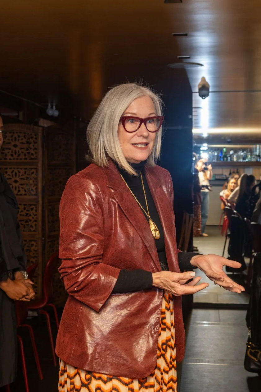 A woman with gray hair, red glasses, and a red leather jacket standing indoors at a social gathering or restaurant. She is speaking and gesturing with her hands.