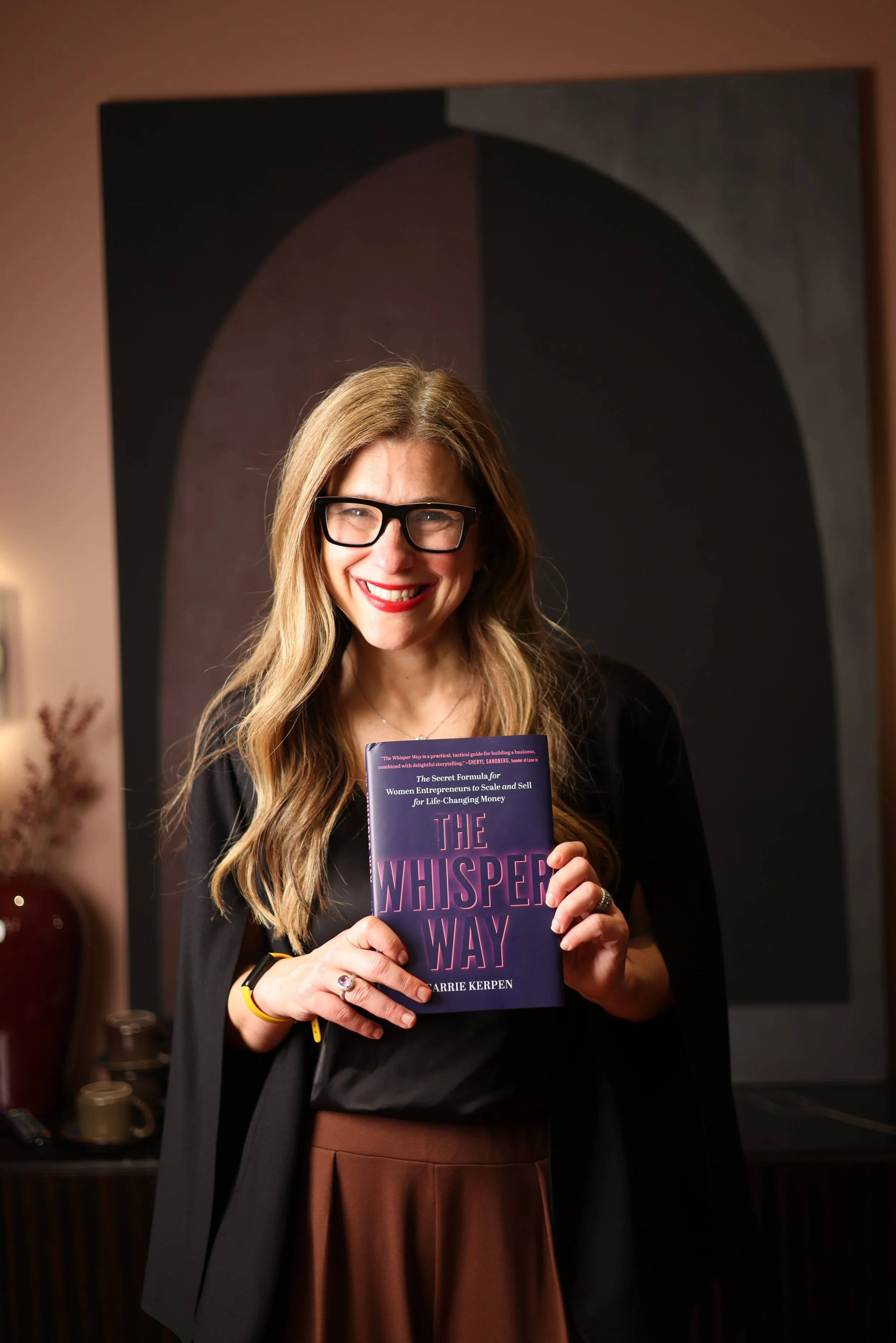 A woman with long blonde hair, wearing glasses and a black top, smiling and holding a book titled 'The Whisper Way' by Carrie Kerpen. She is standing indoors with a dark, abstract artwork on the wall behind her.