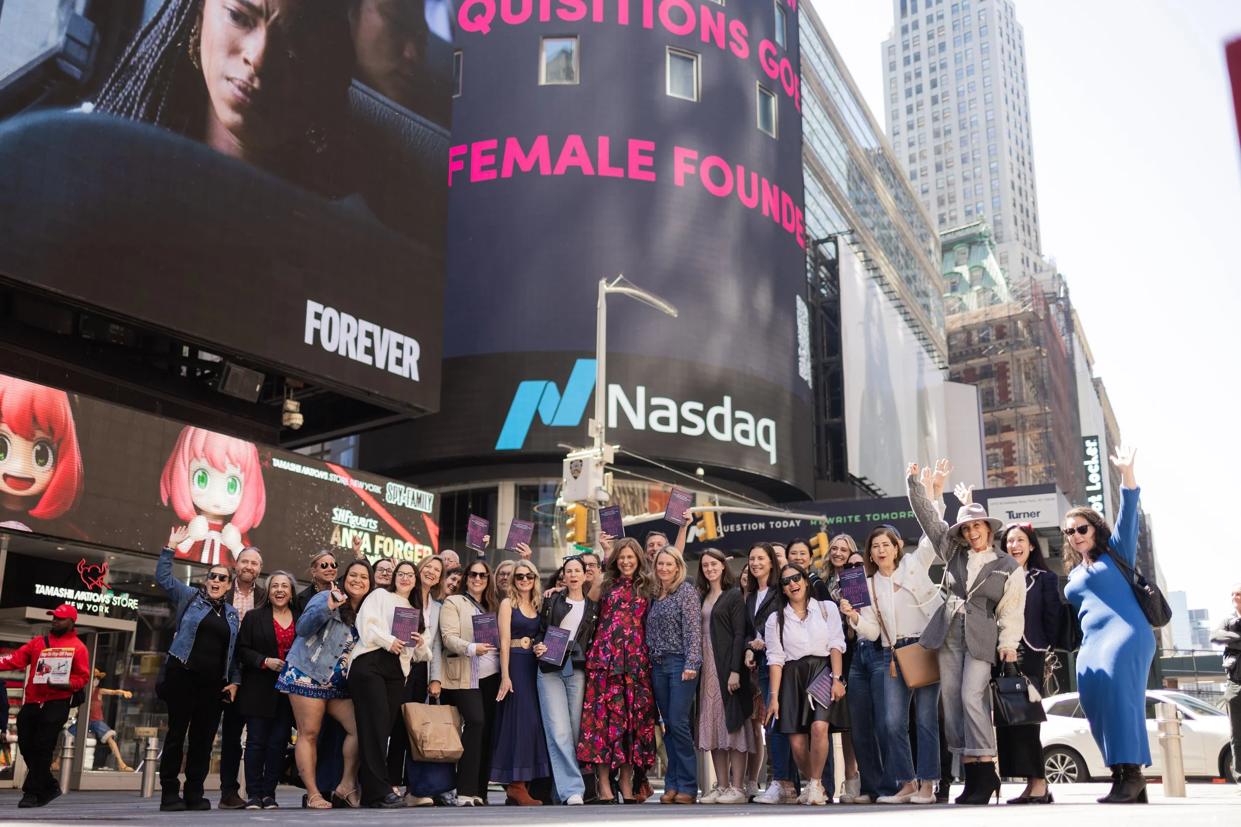 Group of diverse women and men smiling and waving in Times Square, New York City, during a public event, with large digital billboards displaying advertisements and a crowd of pedestrians in the background.