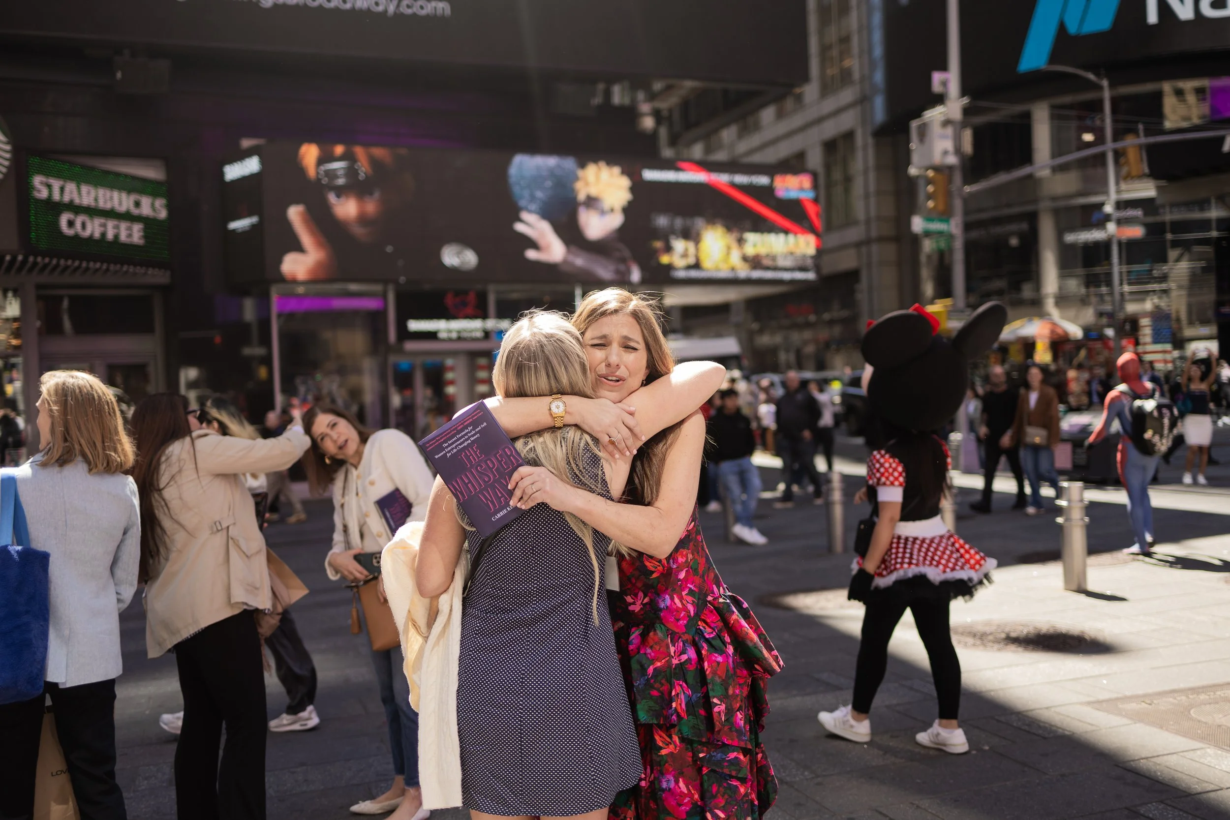 Two women embrace on a busy city street, surrounded by people and bright digital billboards. The woman facing the camera has light brown hair and is holding a book titled 'The Whispering' while crying. The woman with her back to the camera has blonde