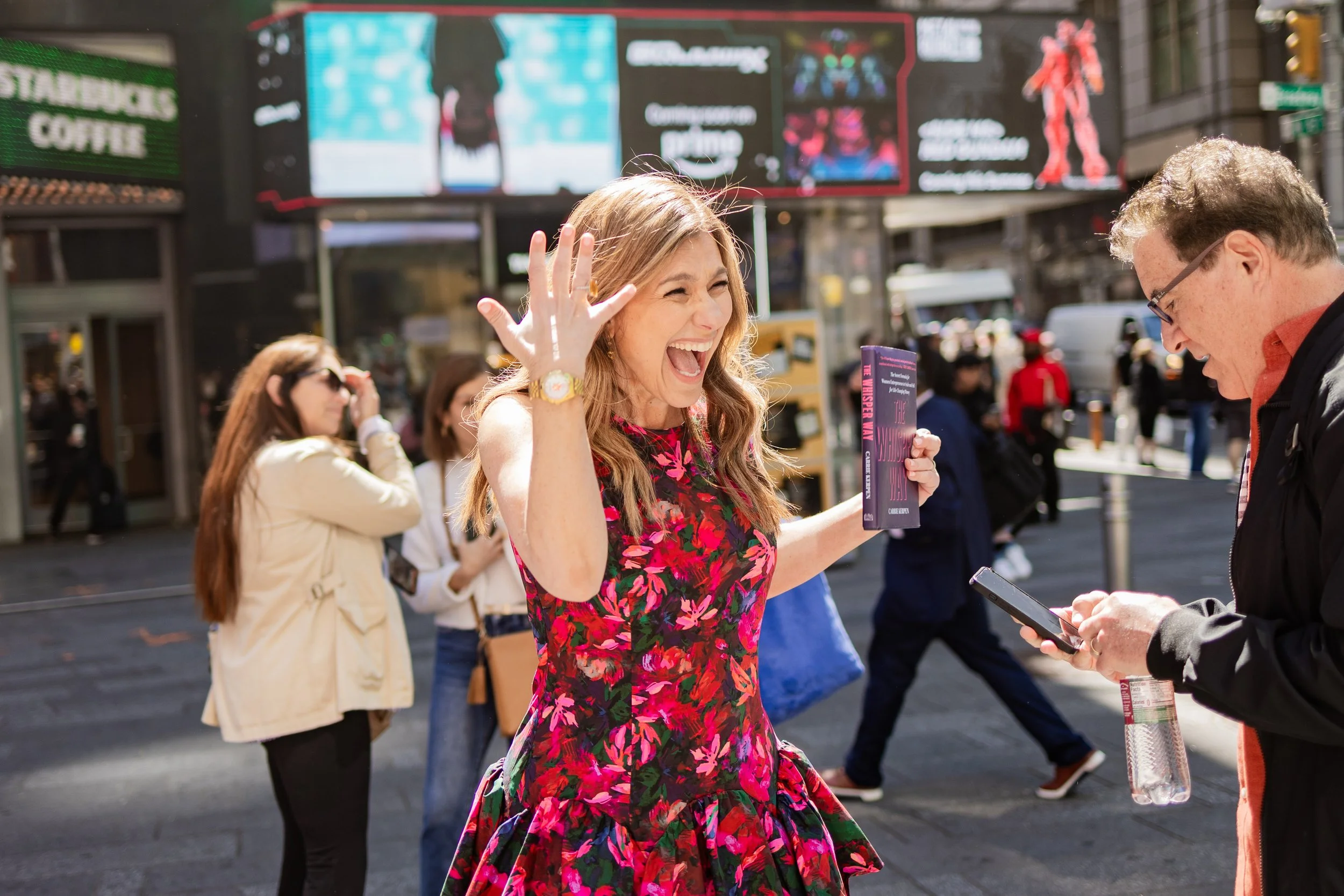 A woman in a pink floral dress laughing and waving her hand while talking to a man with glasses in an urban setting with people and digital billboards in the background.