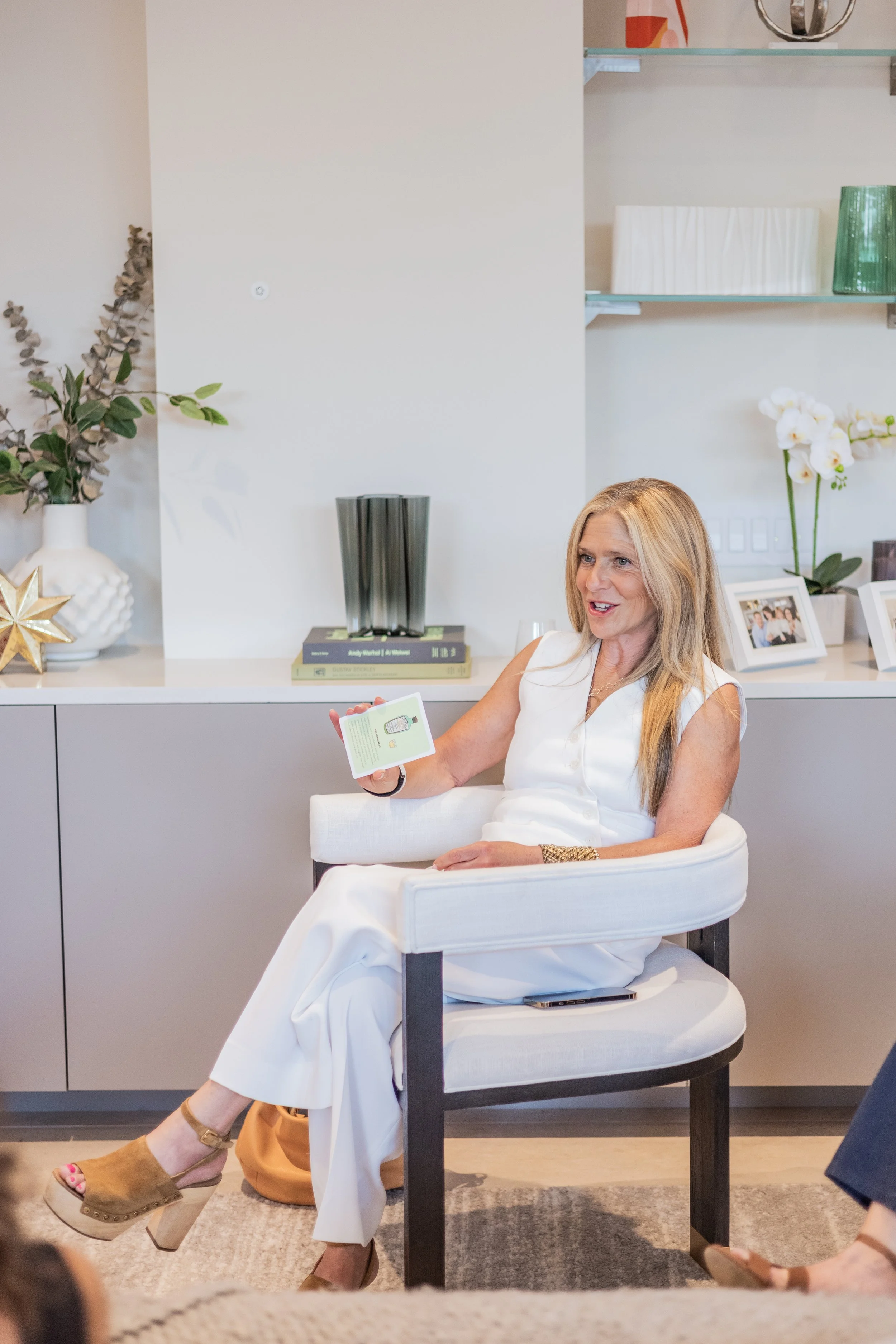 Woman with long blonde hair sitting on a white chair, wearing a white dress, holding a booklet, smiling, in a modern, decorated living room.
