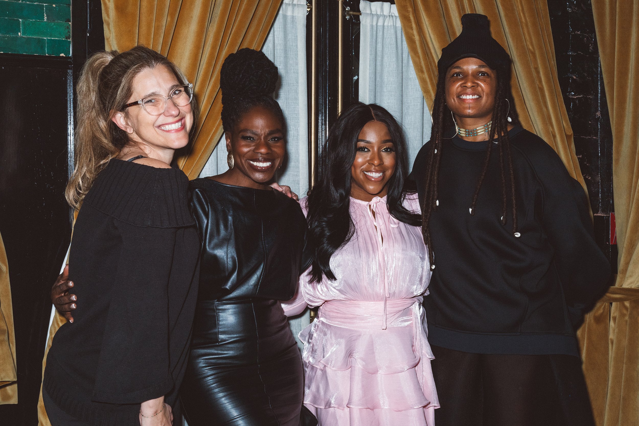 Four women posing together indoors, smiling, with gold and black curtains in the background.