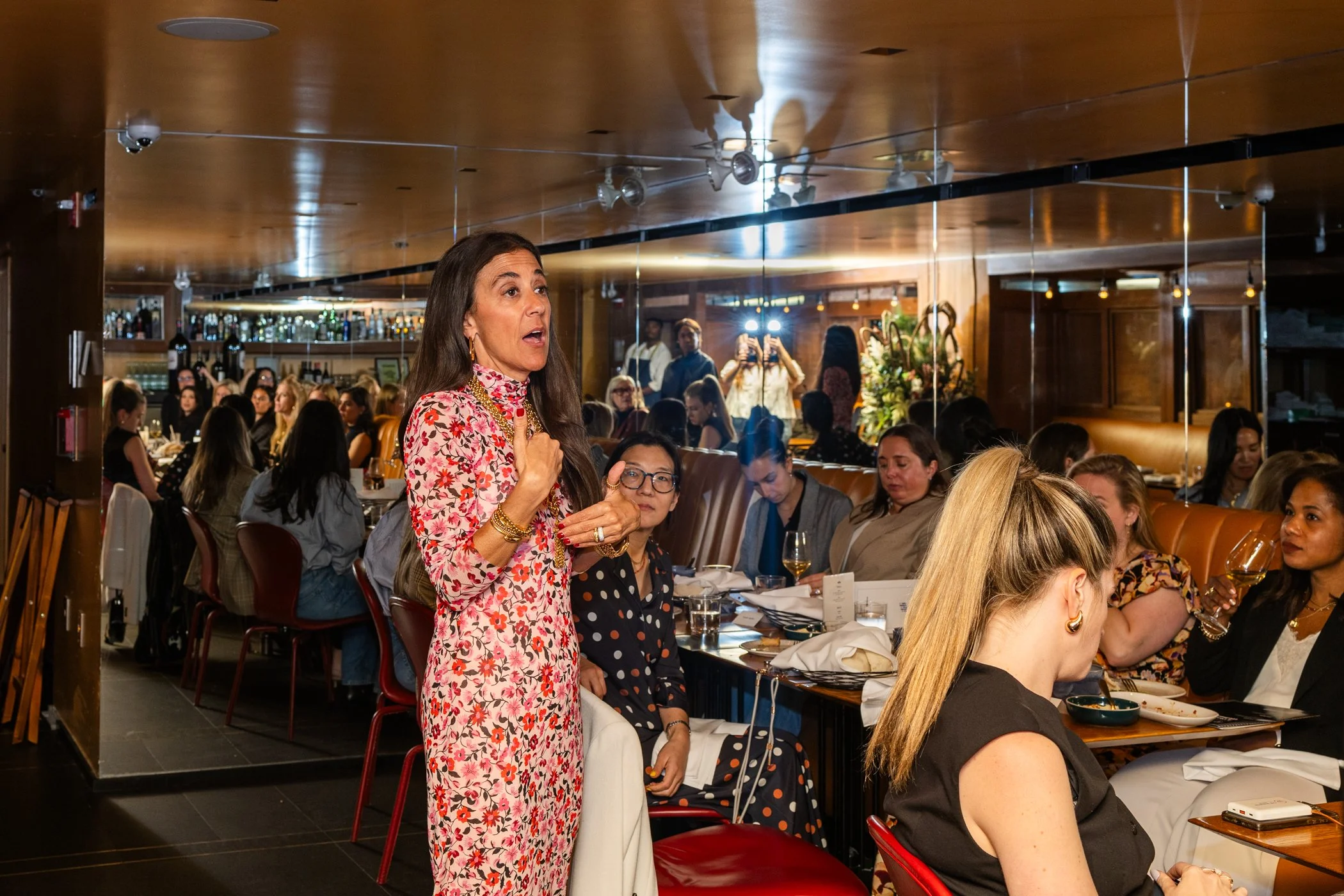 A woman in a pink and red floral dress is speaking at a restaurant gathering, with many people sitting at tables around her, some taking photos and others listening attentively.