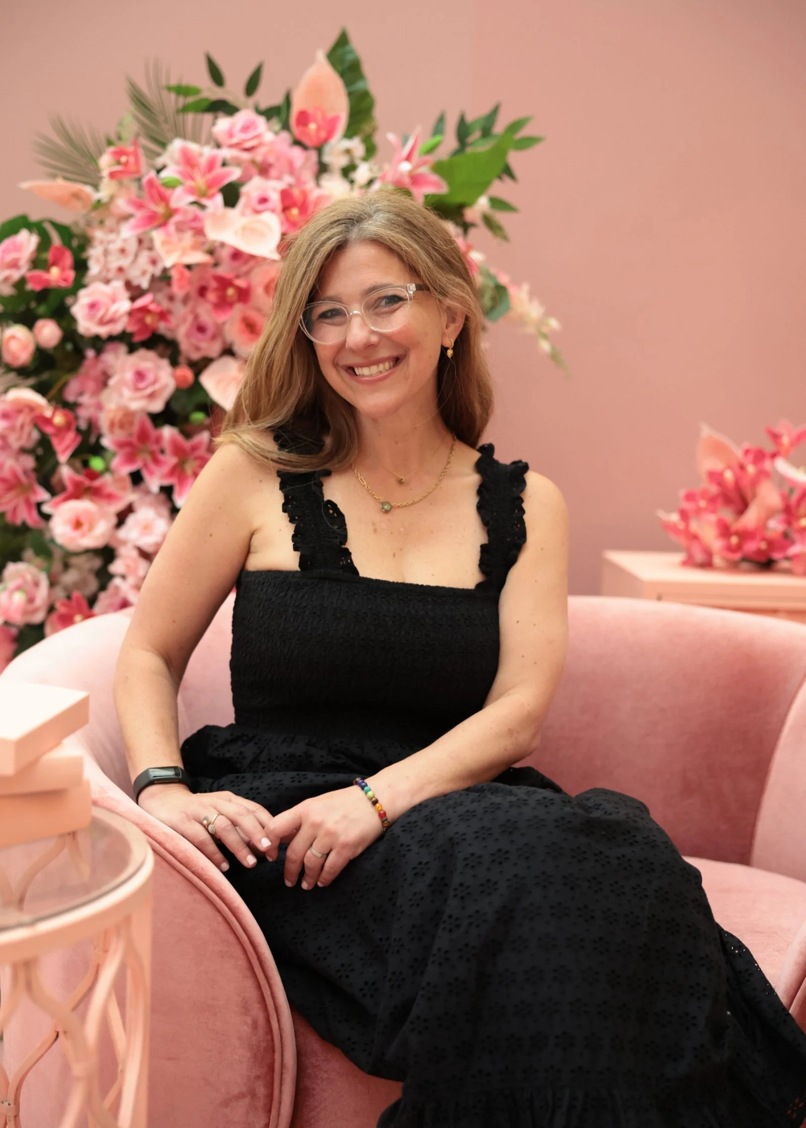 A woman with glasses and long brown hair sitting on a pink velvet couch in front of a large pink floral arrangement, wearing a black dress with lace details, smiling at the camera.