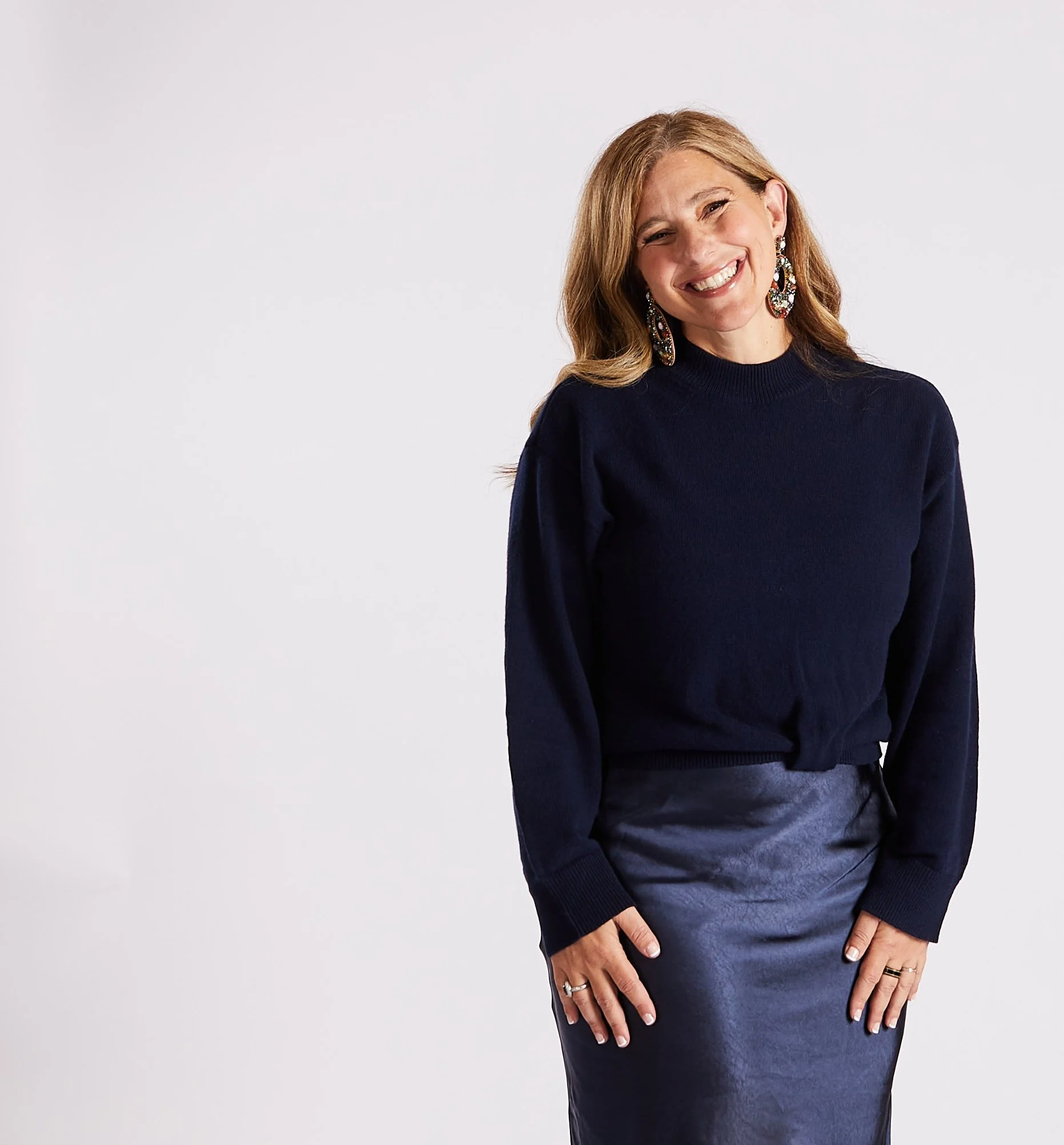 A woman with long, light brown hair and earrings smiling, wearing a dark blue sweater and skirt, posing against a plain white background.