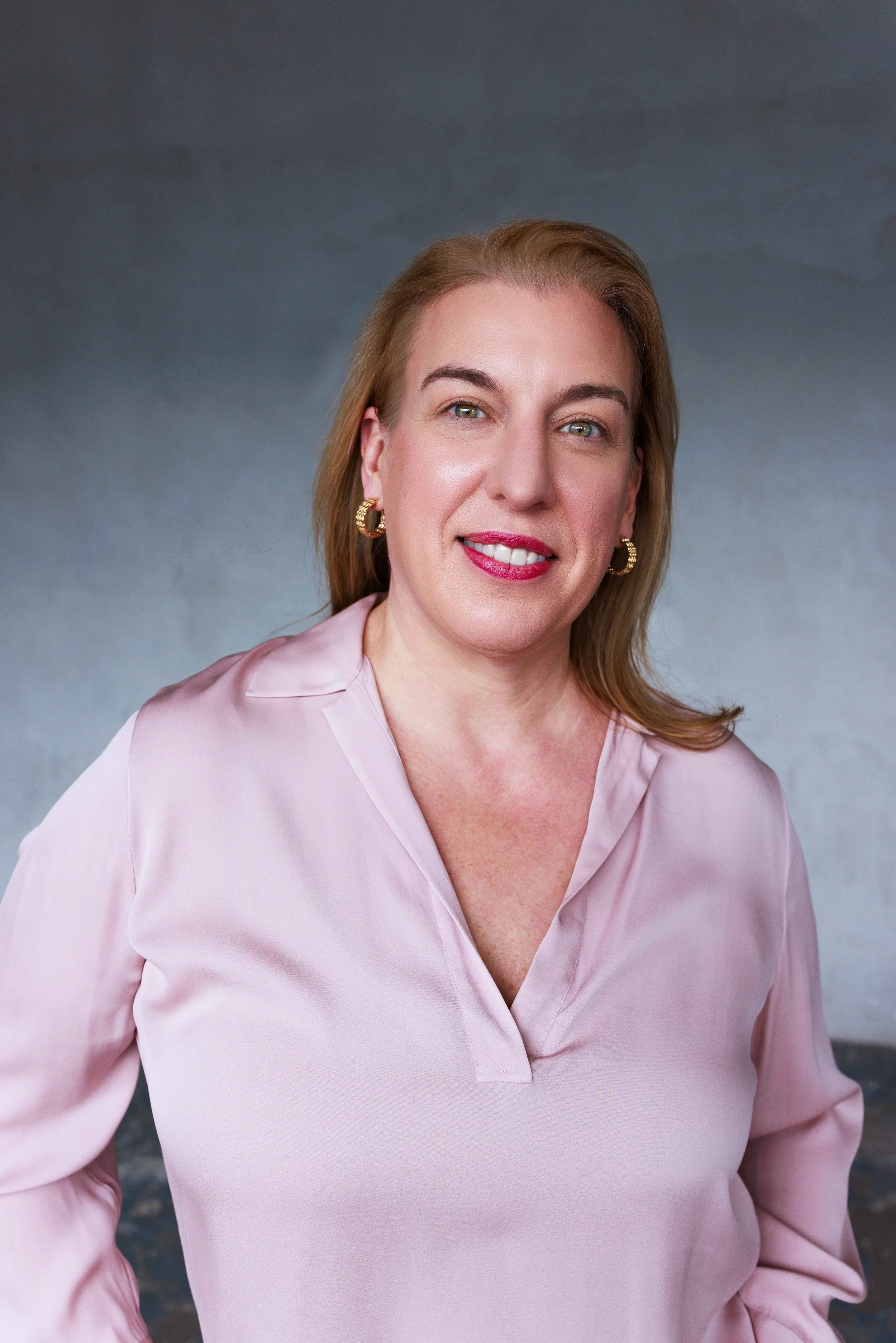 A woman with shoulder-length blonde hair, light skin, and green eyes, wearing gold hoop earrings and a pale pink blouse, stands against a gray background, smiling softly.