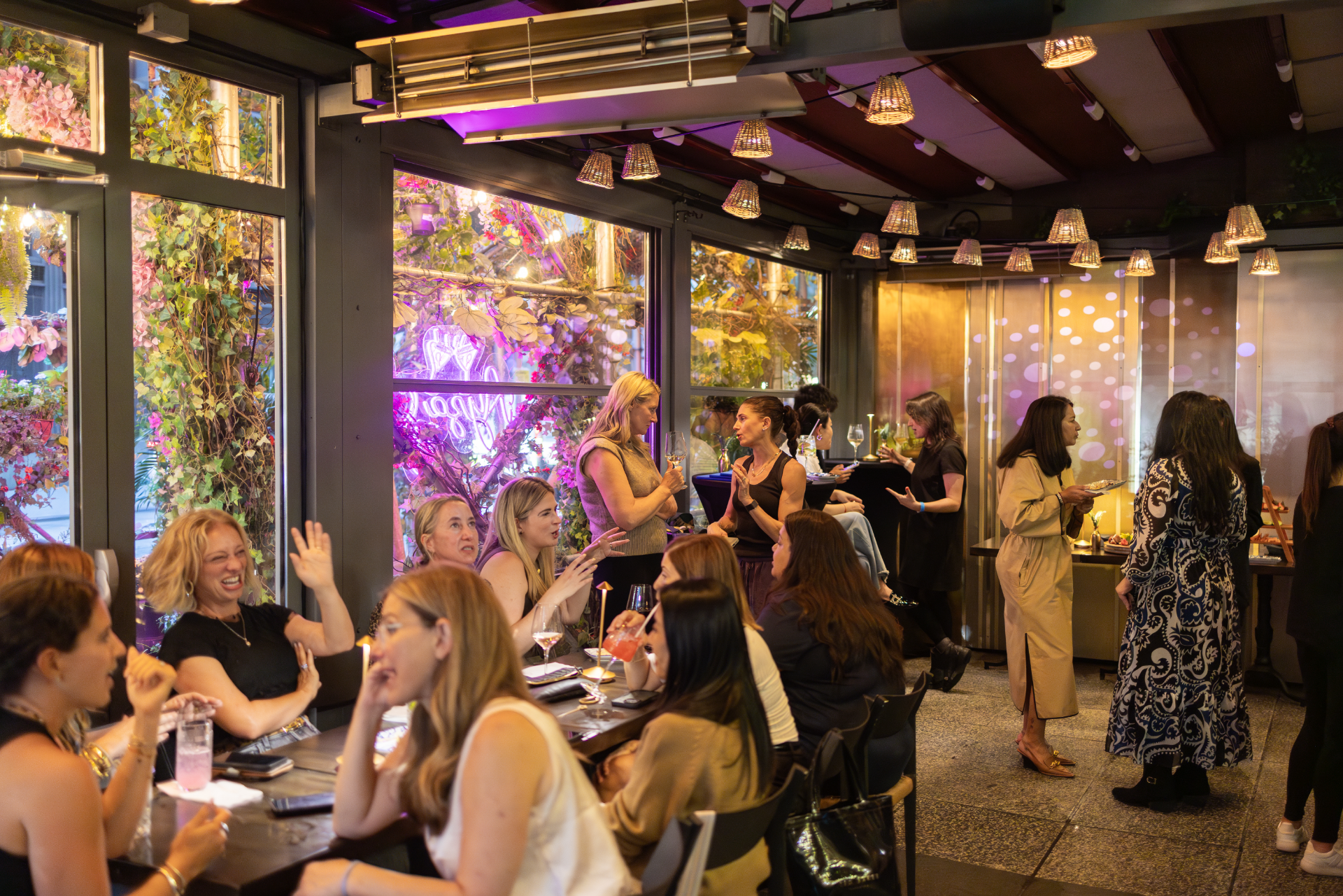 A lively indoor restaurant or bar with a group of predominantly women socializing, some sitting at tables with drinks and food, others standing and engaging in conversation. The decor features large windows with floral arrangements, purple lighting, 