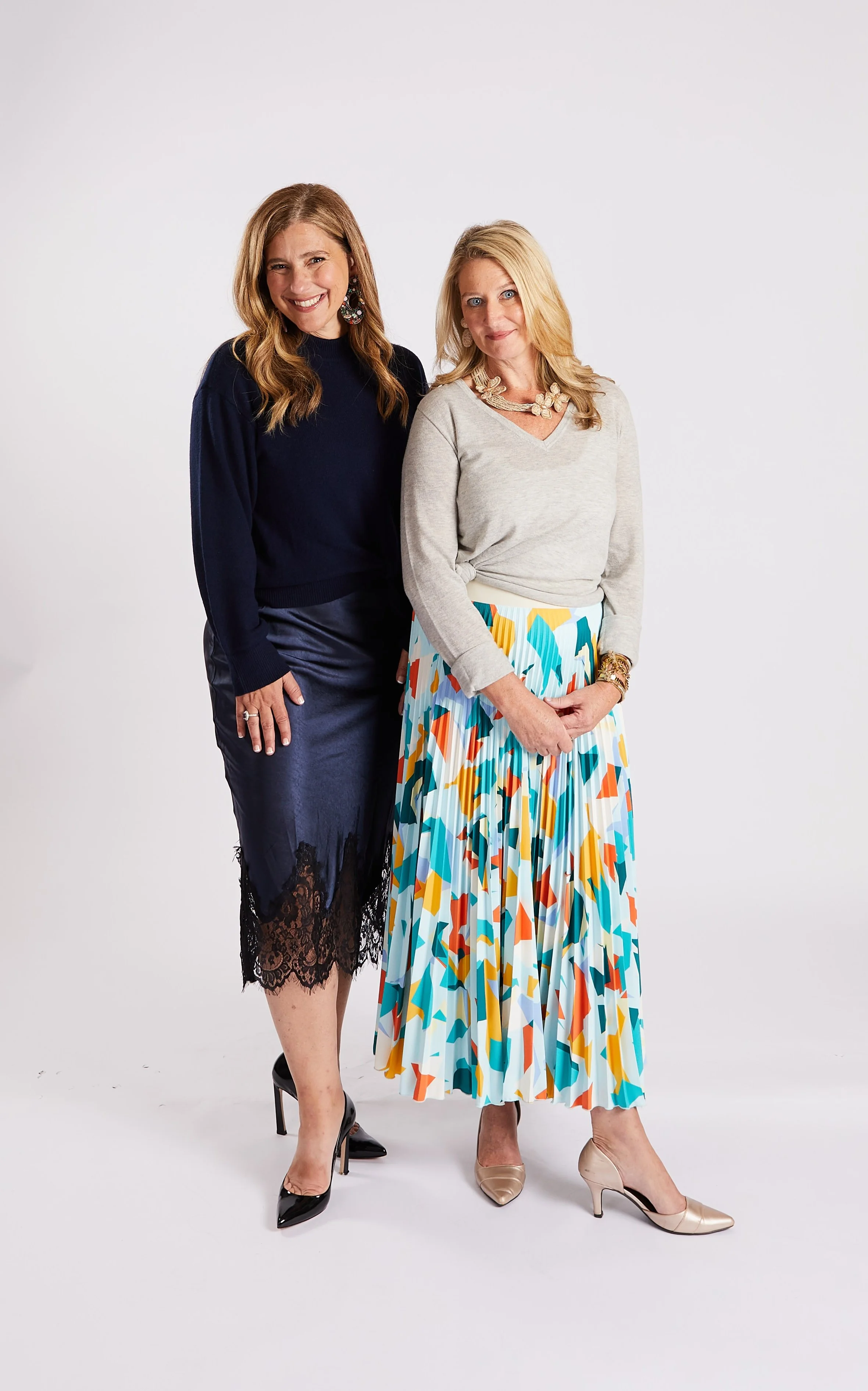 Two women standing together against a plain white background, smiling at the camera. One woman has long, wavy brown hair and is wearing a navy blue top with a black lace skirt and black high heels. The other woman has long, straight blonde hair and is wearing a light gray sweater, a colorful patterned pleated skirt, and beige high heels.