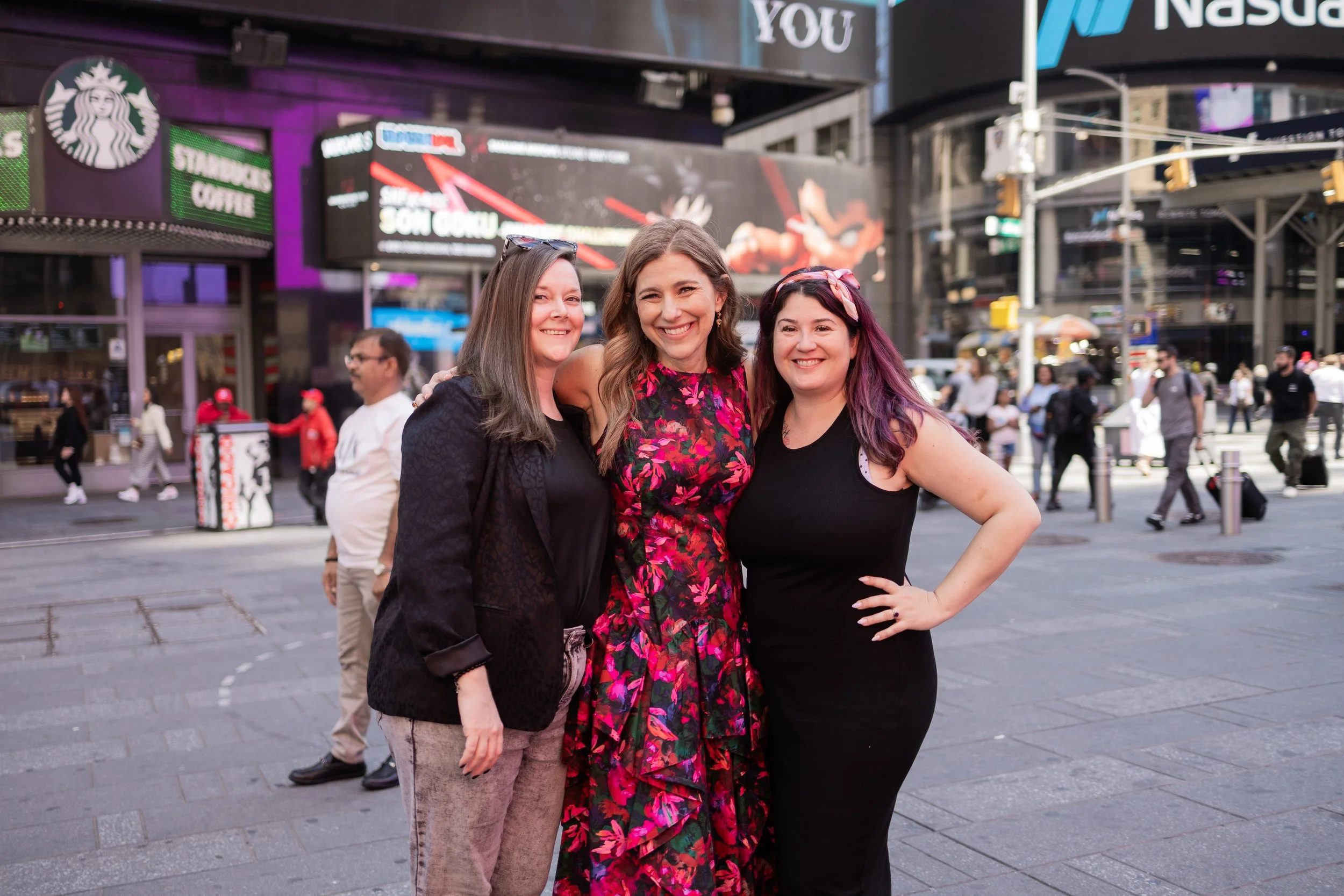 Three women smiling in Times Square, New York City, with bright digital billboards and crowds in the background.