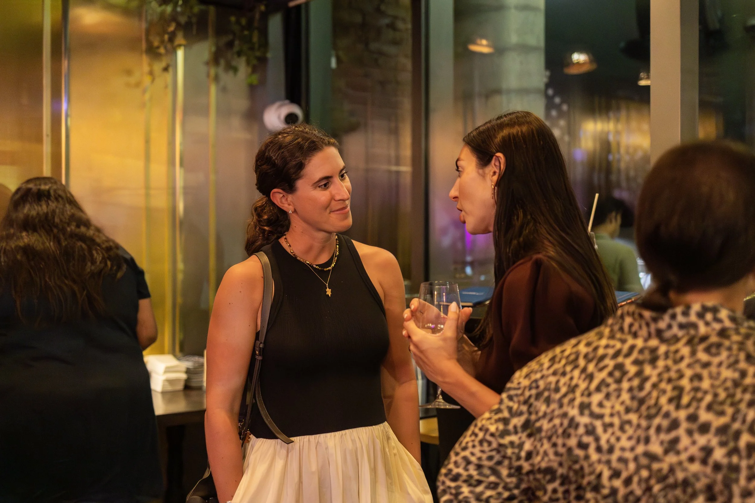 Two women are engaged in conversation at a social gathering, with one holding a glass of wine. The woman on the left is wearing a sleeveless black top, a beige skirt, and a necklace, while the woman on the right is in a brown top. The background show