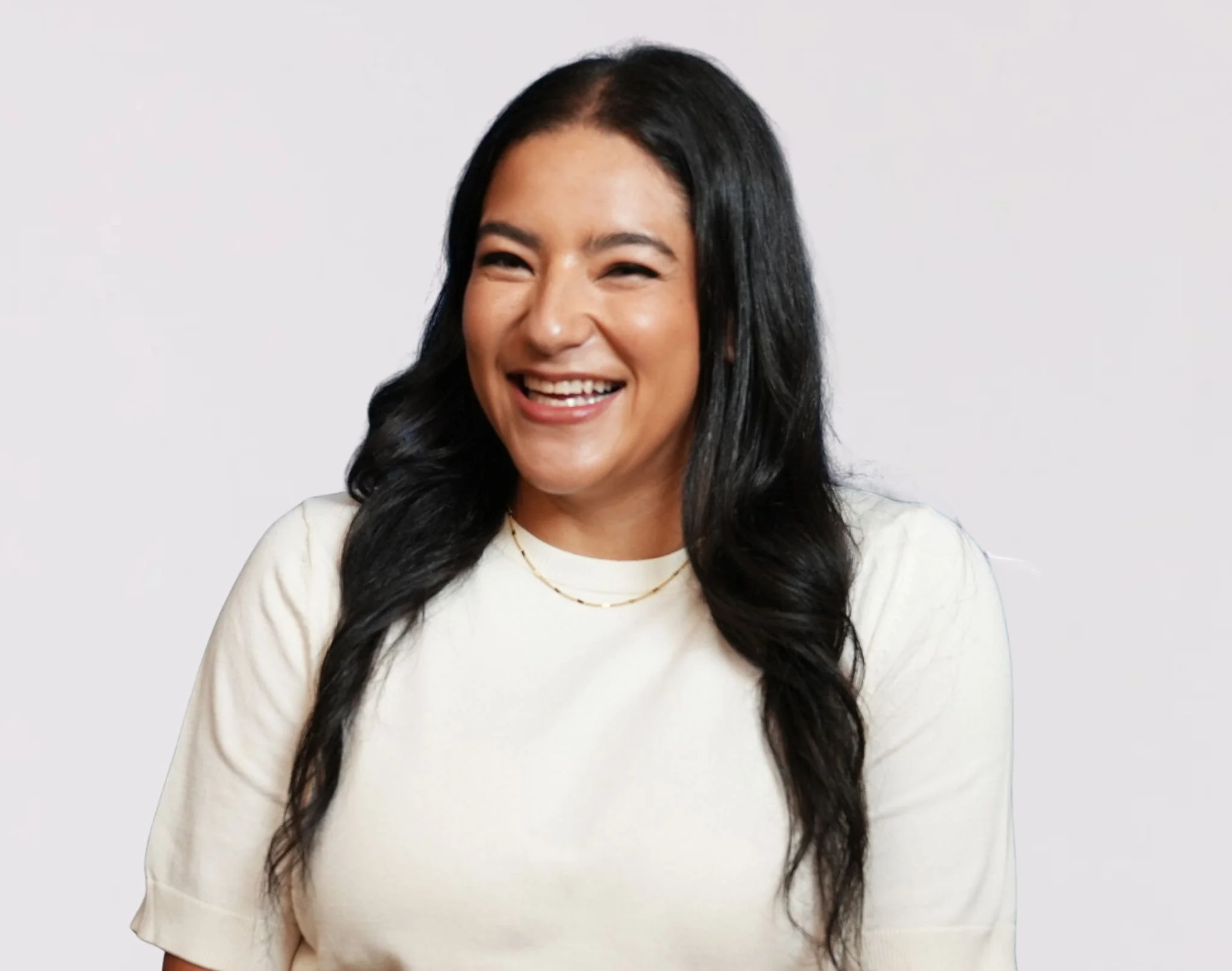 A woman with long dark hair and a bright smile wearing a white top and gold necklace, standing against a plain white background.