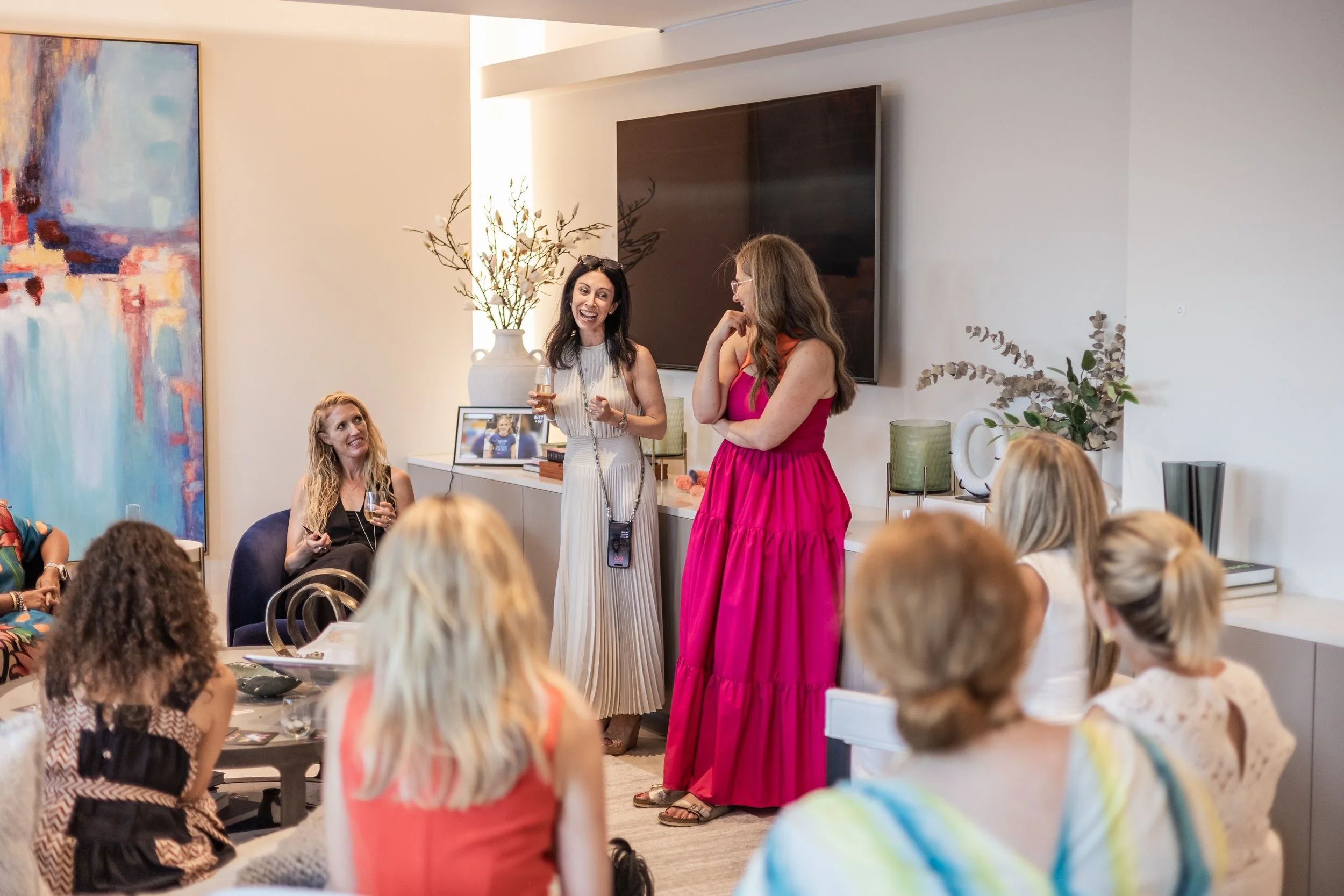 A group of women in a living room, some sitting and some standing, engaged in conversation. The woman standing in the center is holding a glass and speaking, with others listening and smiling. The room has modern decor with a large abstract painting on the wall, plants, and a television.