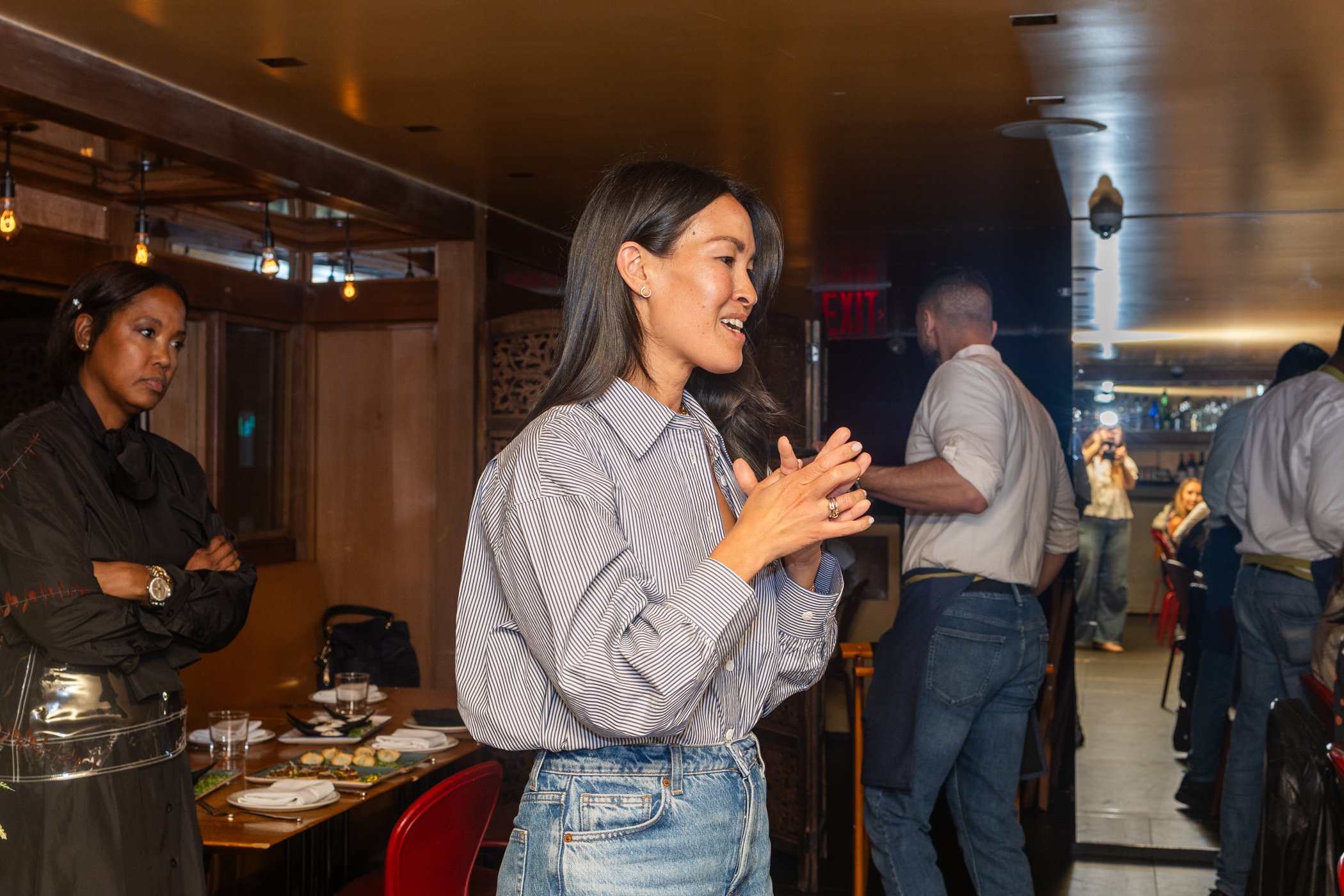 A woman with long dark hair standing in a restaurant, hands clasped together, smiling and engaged in conversation. Several other people are visible in the background, some looking at her.