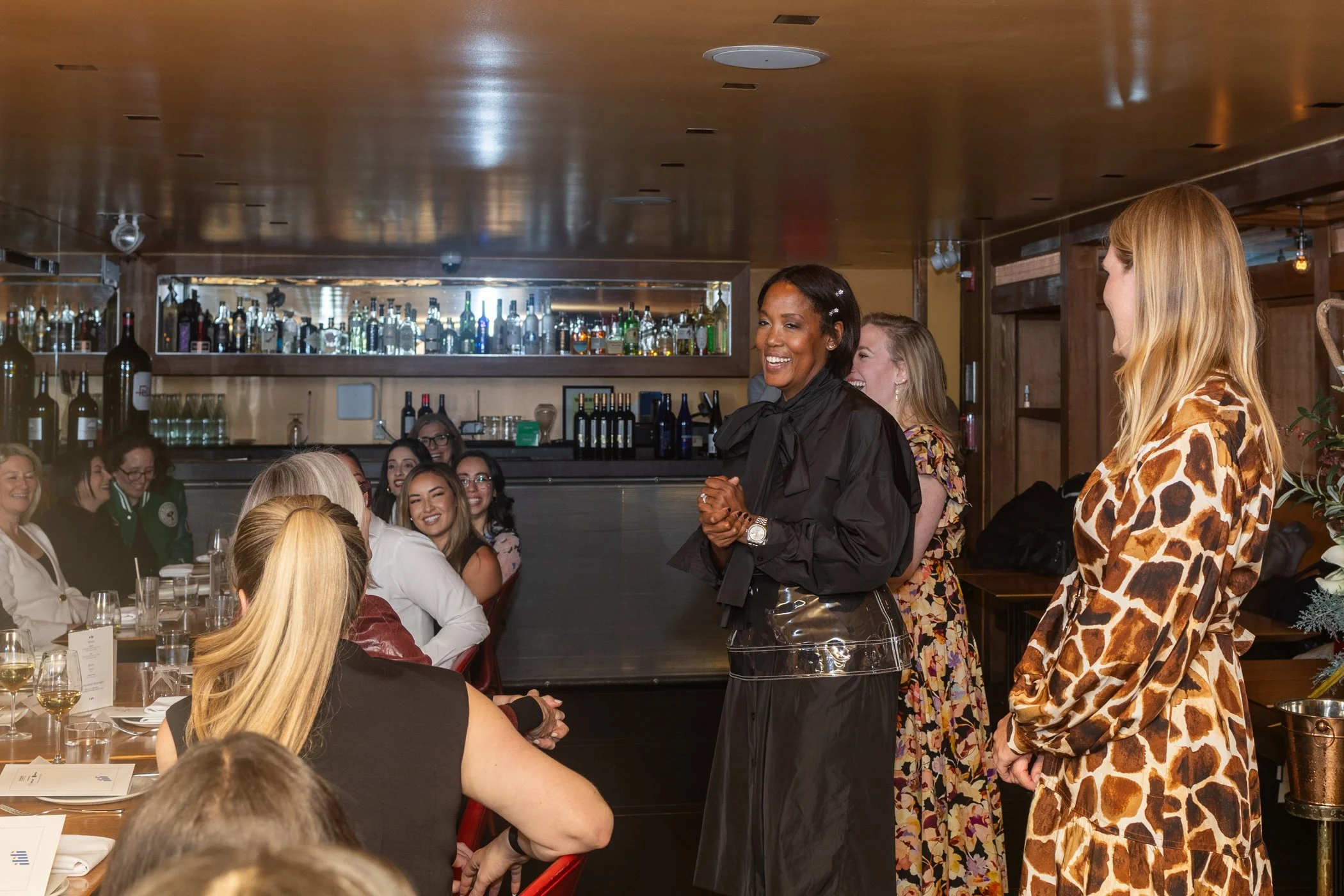 Group of women sitting at a restaurant table with drinks, while a woman in a black outfit and floral dress speaks to a woman in a giraffe-print dress, with a bar in the background.