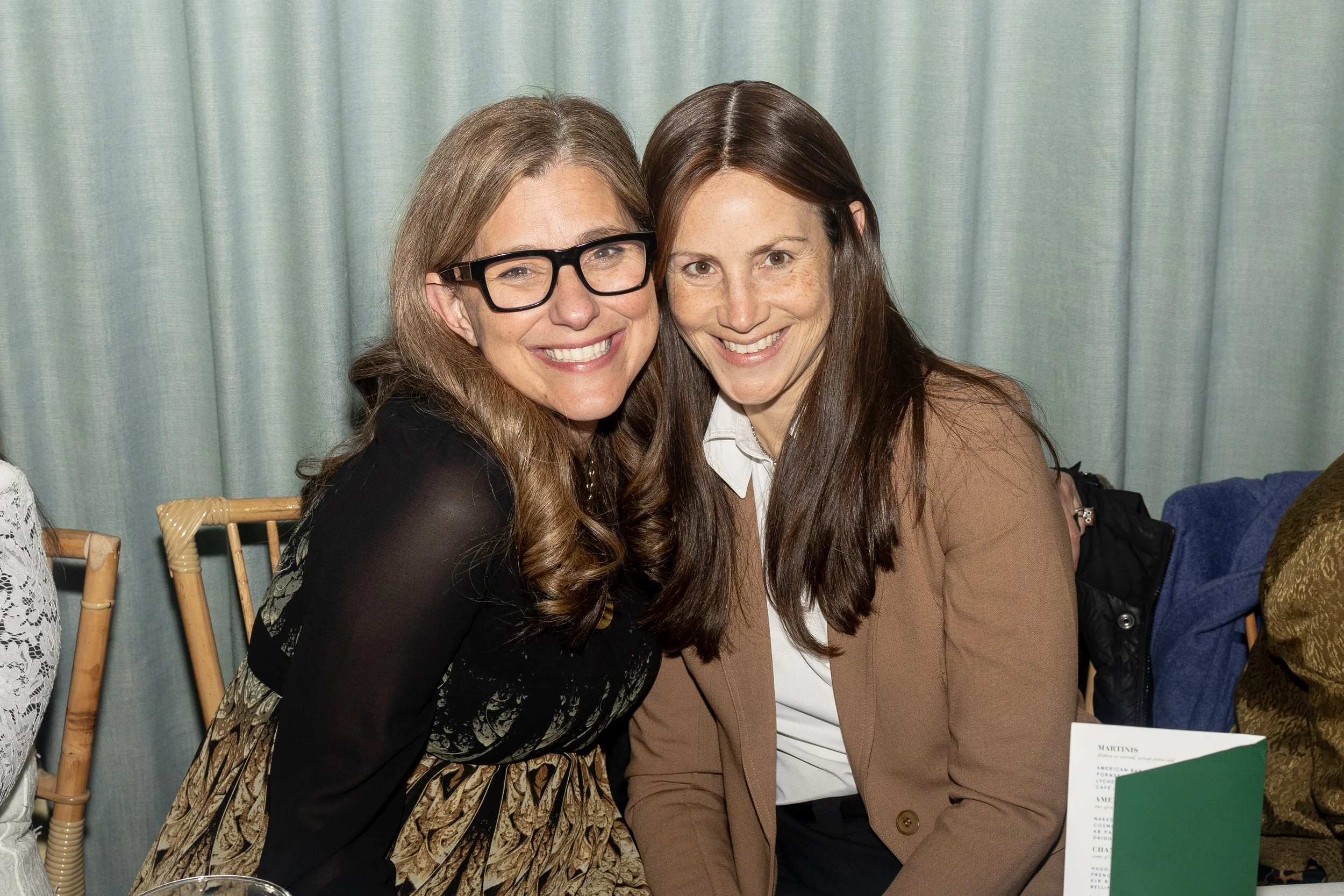 Two women smiling and sitting closely together at an event. One woman has long brown hair and freckles, wearing a beige blazer. The other woman has long light brown hair, glasses, and is wearing a black dress with a patterned skirt.