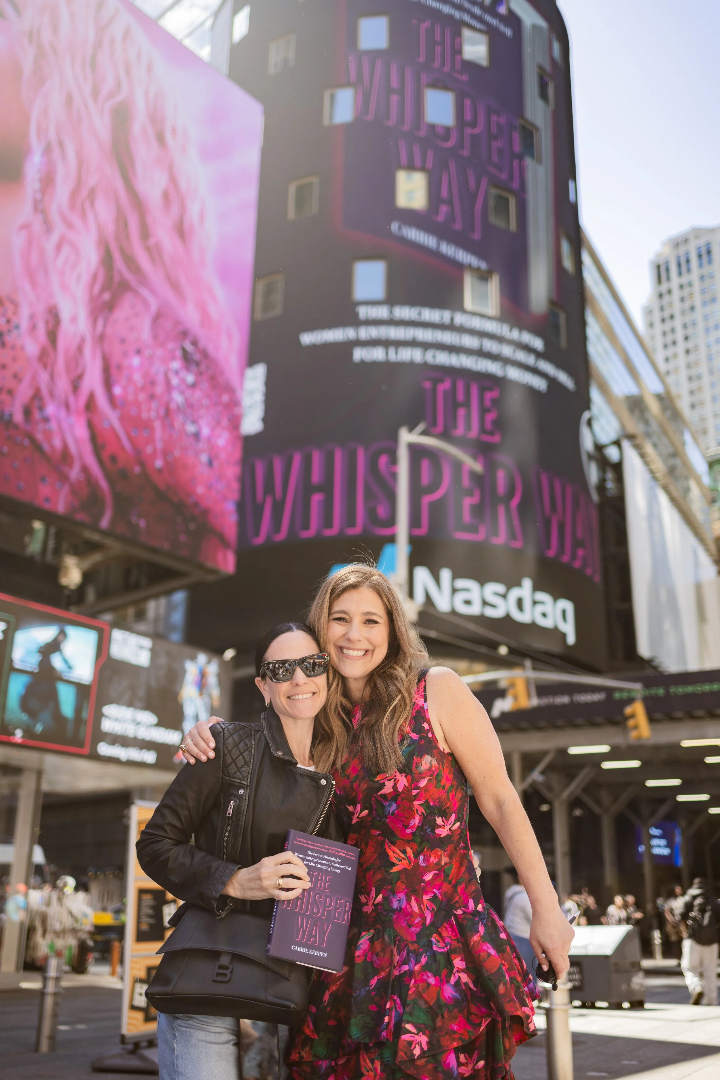 Two women standing together outdoors in Times Square, New York City, smiling at the camera. One woman is holding a book titled 'The Whisper Way' and the other is holding a phone. Behind them, digital billboards and city buildings are visible.